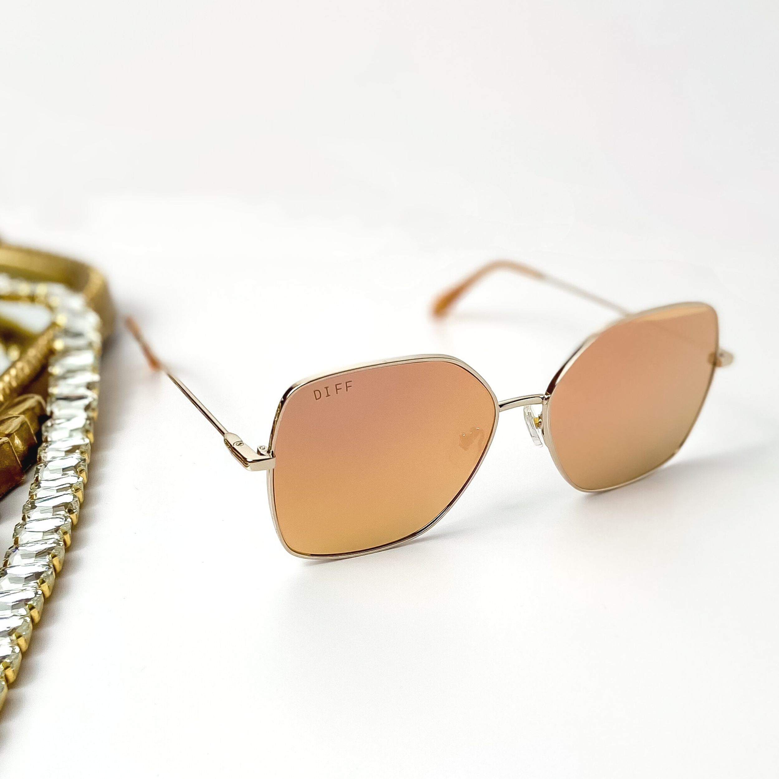 A pair of gold tone sunglasses with peach orange mirror lenses. Pictured on a white background with gold jewelry.
