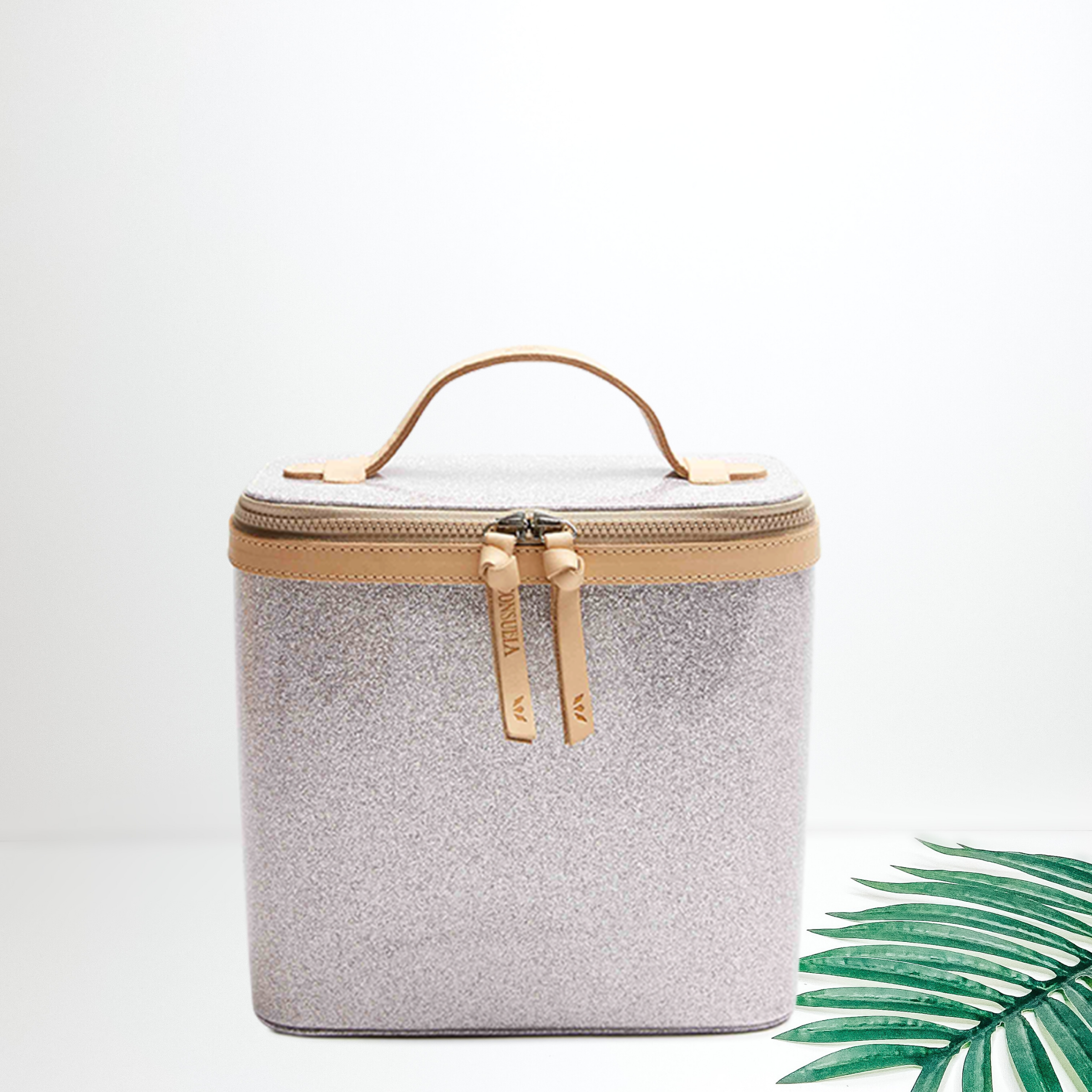 A light lilac glitter tall train case with leather details. Pictured on white background with a palm leaf and black sunglasses.