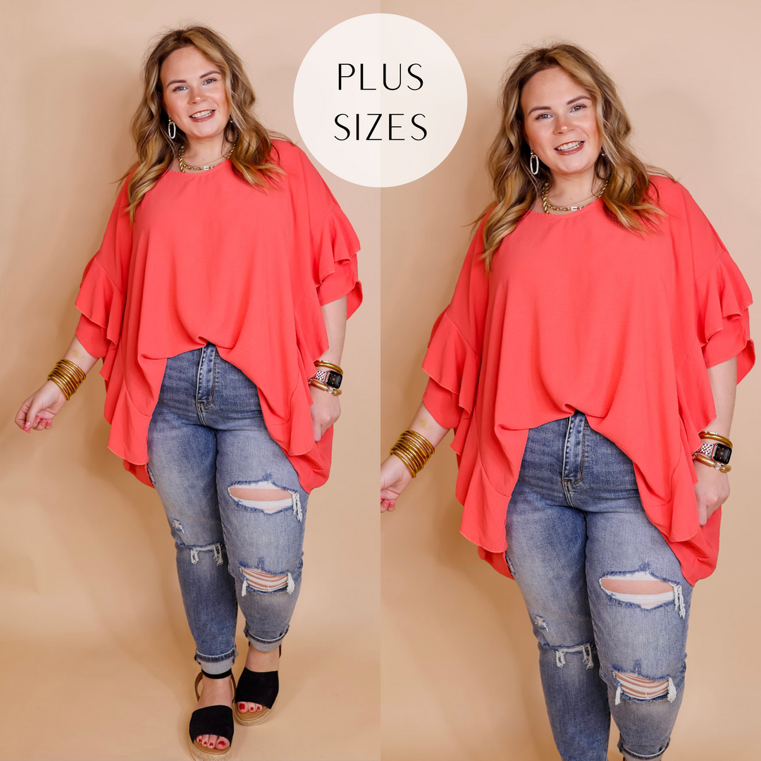 Model is wearing a coral pink poncho top with ruffle sleeves and ruffles down the side. Model has it paired with distressed skinny jeans, black sandals, and gold jewelry.