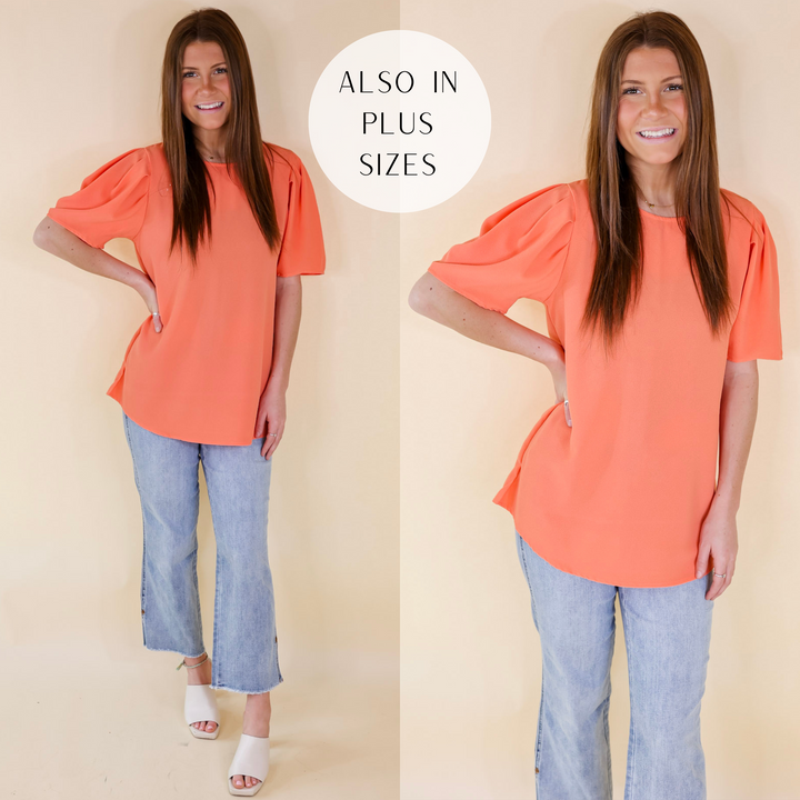Model is wearing a short sleeve top with pleated detail on the shoulder. Model has this orange top paired with light wash jeans, ivory heels, and gold jewelry.
