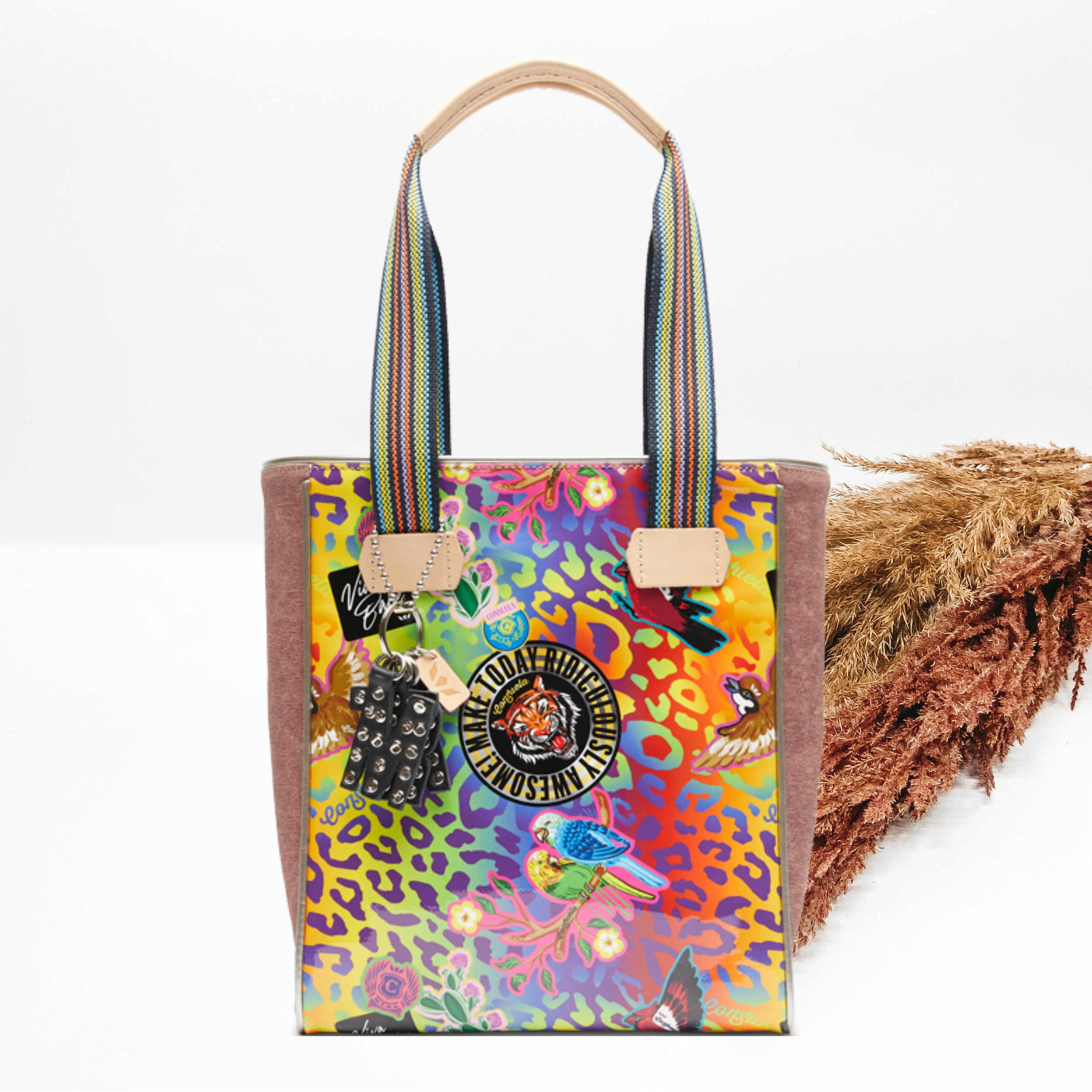 Pictured is a tote bag with striped straps and a black charm hanging on one of the straps. This tote has a multicolored leopard print design that includes different pictures randomly throughout and includes light mauve fabric sides. This tote is pictured on a white background with pompous grass on the right side of the picture.