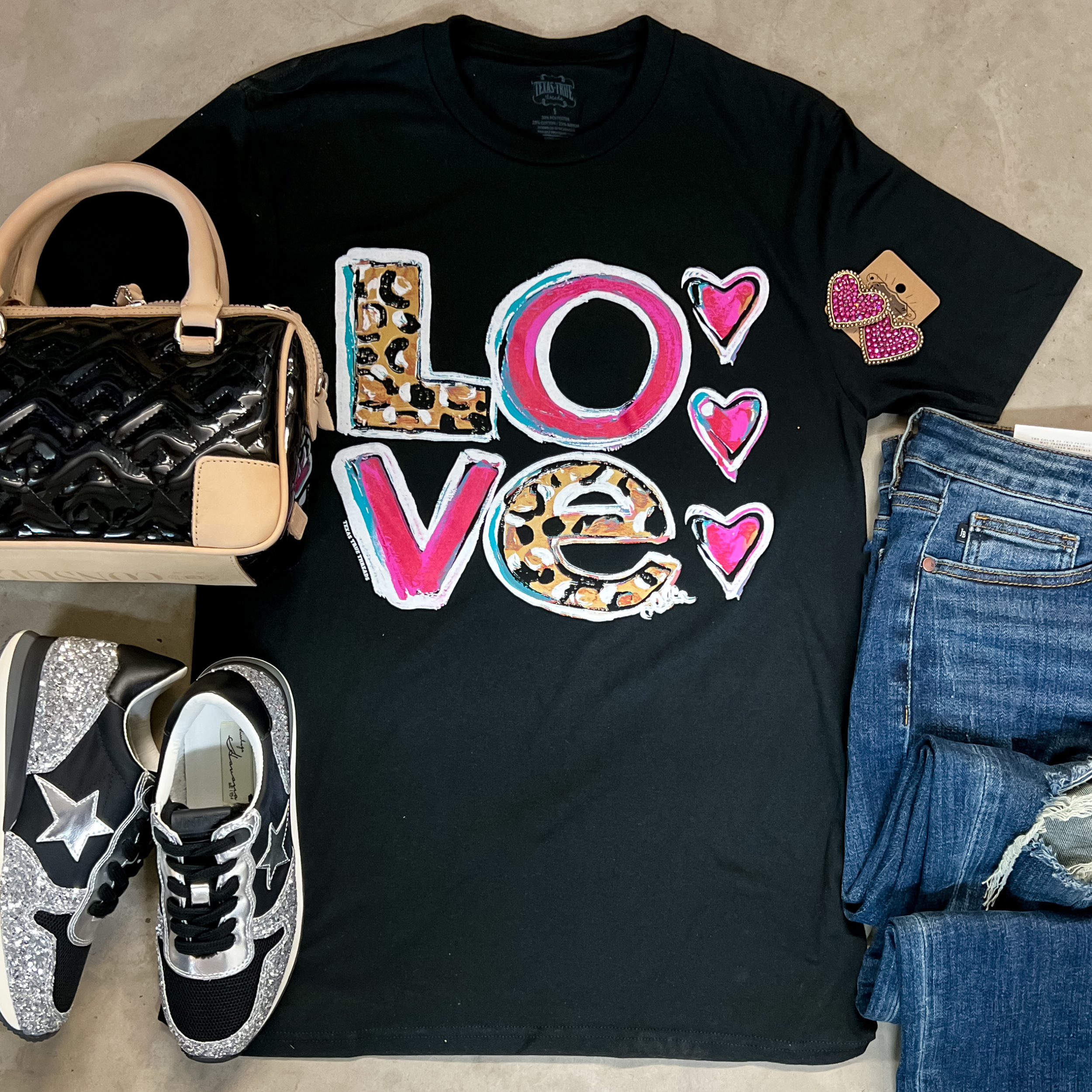 A black tee shirt with short sleeves, a crew neckline, and a graphic that says "LOVE" with pink, blue, and leopard print letters. It is pictured with a black purse, black and silver sneakers, and distressed jeans.
