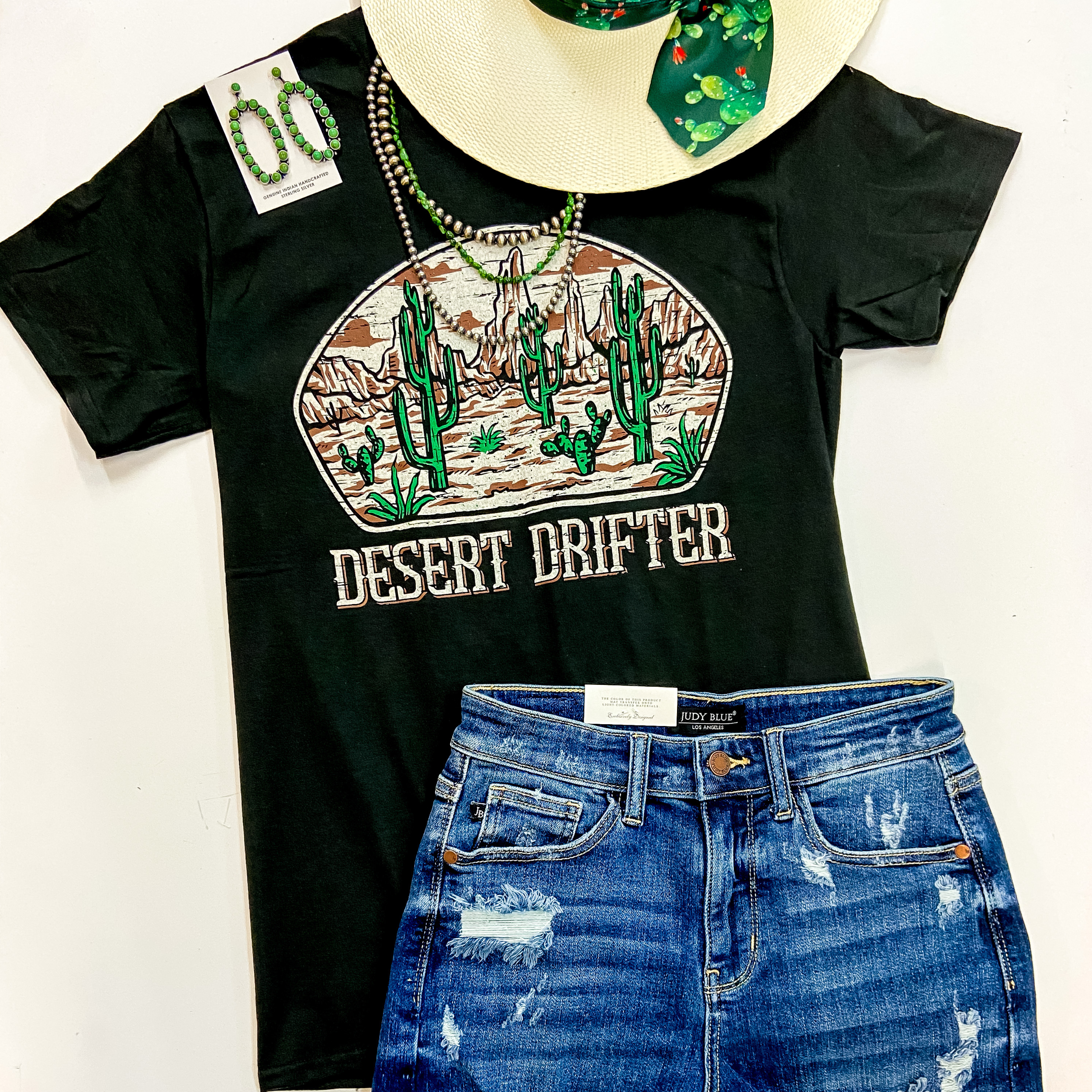 A black short sleeve tee shirt with a desert graphic of mountains and cactus with the phrase "Desert Drifter" written underneath. This tee is pictured on a white background with denim shorts, sterling silver and turquoise jewelry, and a straw hat.