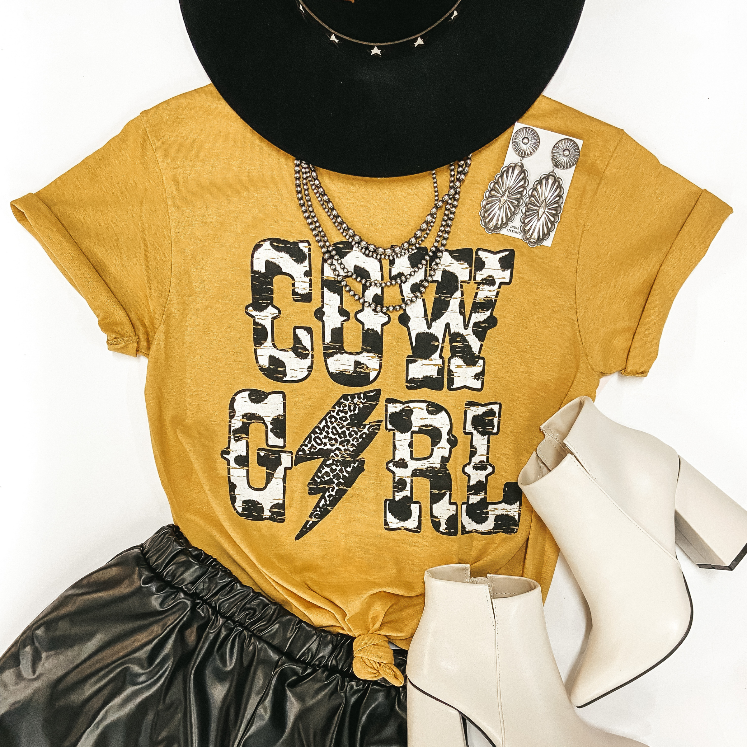 A mustard yellow graphic tee with cuffed sleeves and a knotted front. The tee shirt has a graphic that says "Cow Girl" in cow print with a leopard print lightning bolt in place of the "i" in "girl." Pictured on white background with black hat, black faux leather shorts, ivory booties, and sterling silver jewelry.