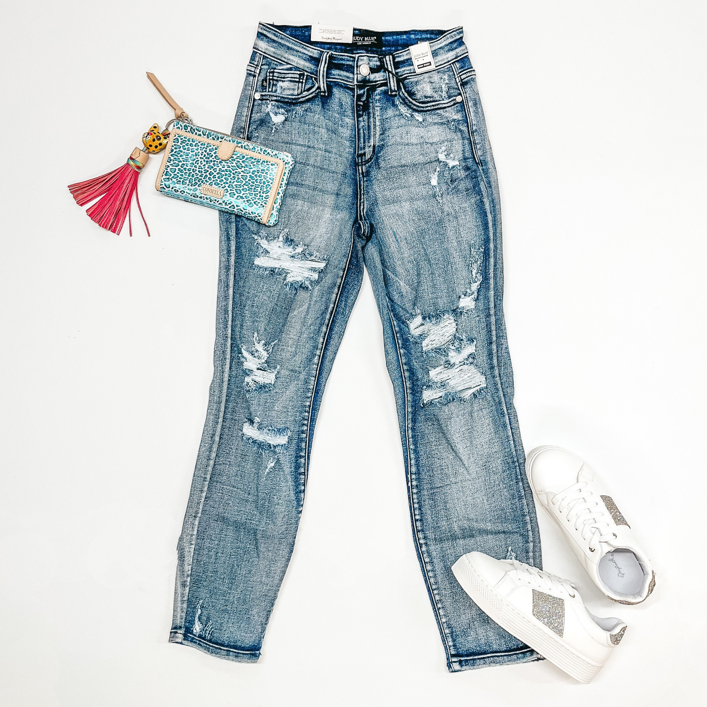 A pair of distressed boyfriend jeans in light wash. Pictured on a white background with white sneakers, a leopard print wallet and keychain.