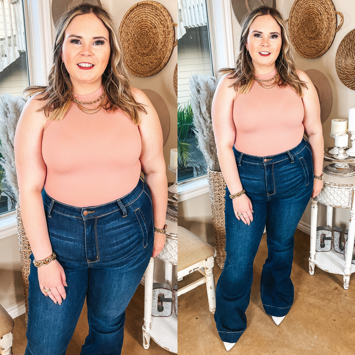 Model is wearing a high neck tank top bodysuit that is blush pink. Model has it paired with dark wash trouser jeans, and gold jewelry.