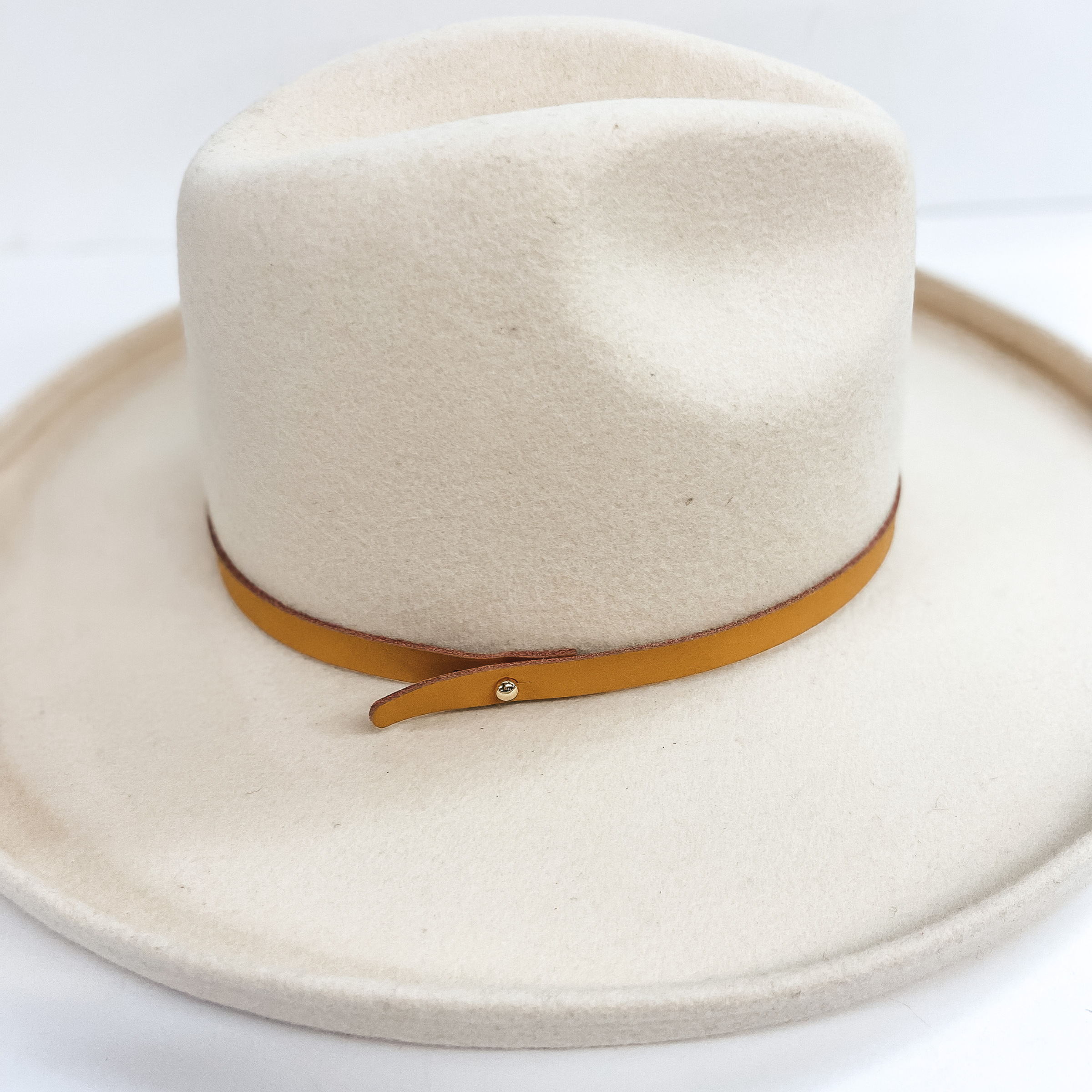 A camel brown thin leather hatband that has a gold pin. Pictured on an ivory felt hat.