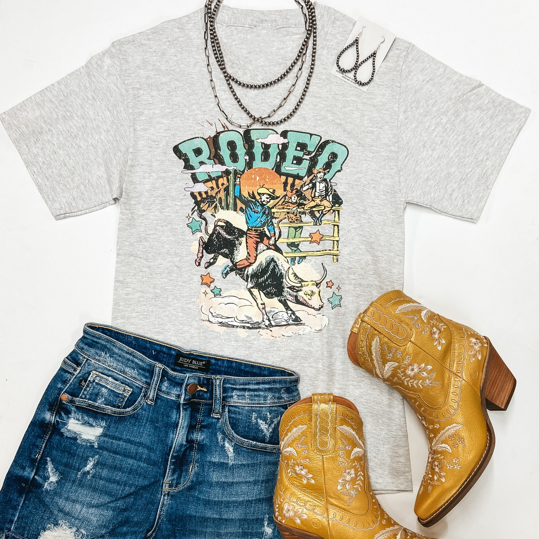 A heather grey graphic tee with the graphic of a cowboy on a bucking bull that says "Rodeo Western." This tee is pictured on a white background with yellow boots, denim shorts, and genuine Navajo jewelry.
