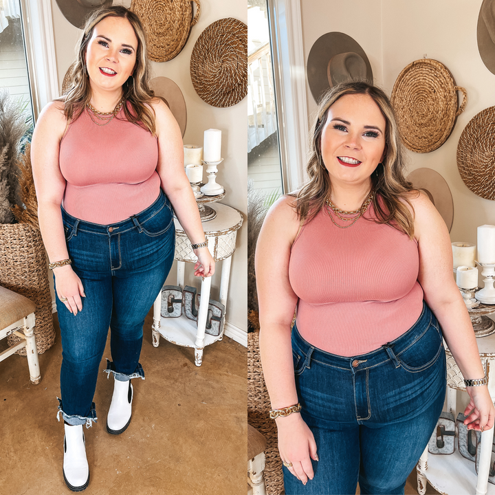 Model is wearing a high neck tank top body suit that is pink. Model has it paired dark wash skinny jeans, white booties, and gold jewelry.