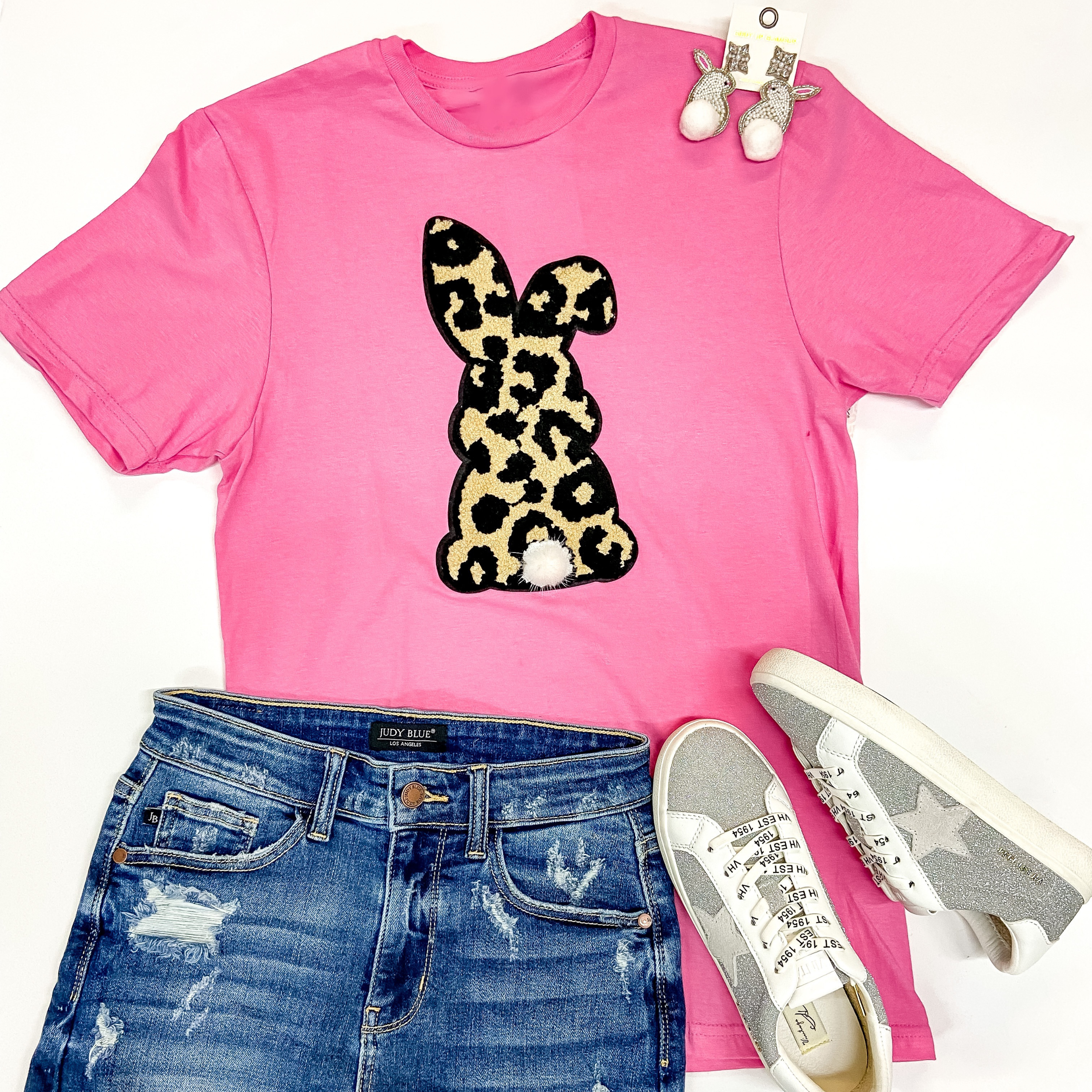  A pink short sleeve tee shirt with a crew neckline and a chenille patch that is leopard print in the shape of a bunny. This tee is pictured on a white background with silver sneakers, bunny shaped earrings, and denim shorts.