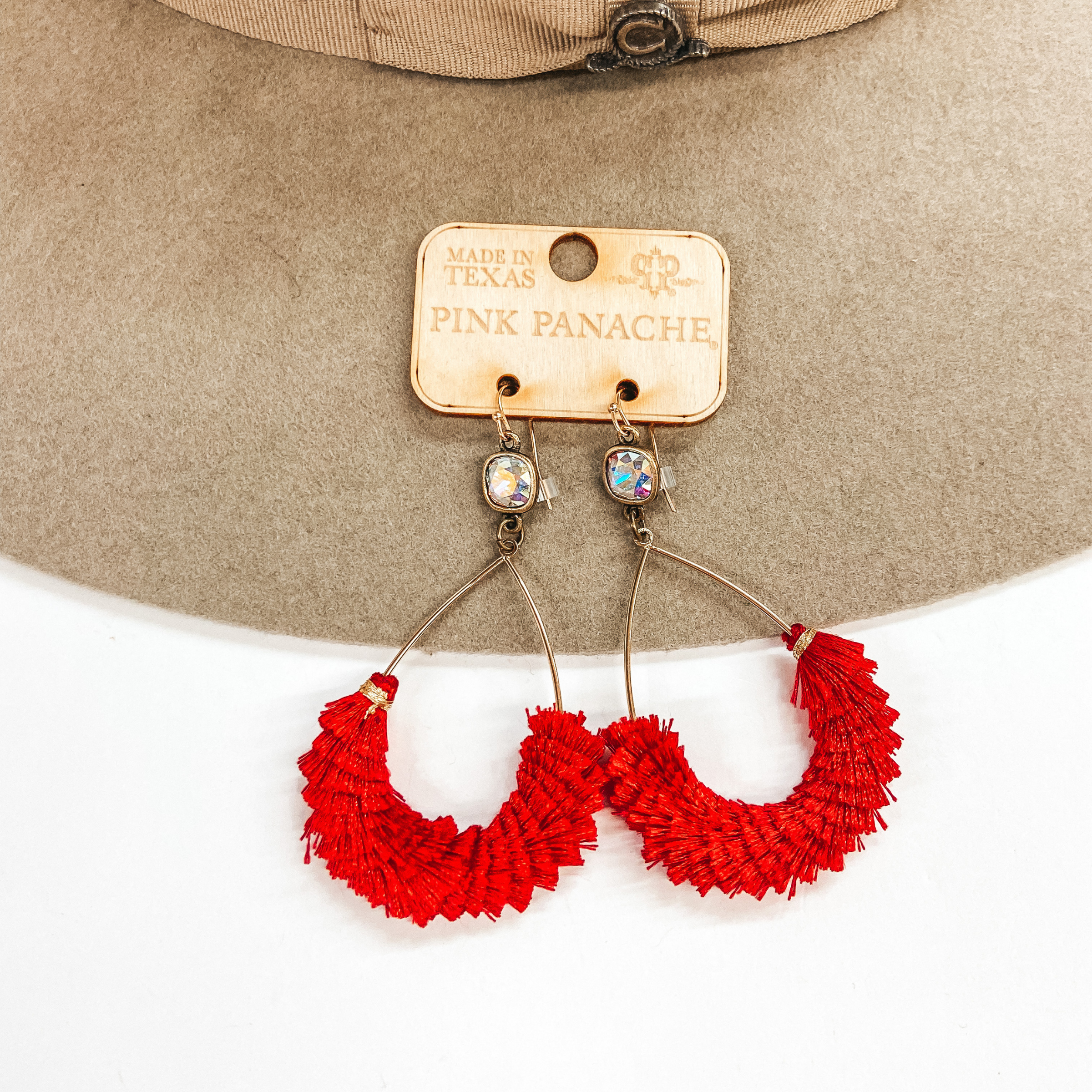 A pair of gold teardrop earrings with an AB cushion cut crystal and red fringe detailing. Pictured on white background with a beige hat.