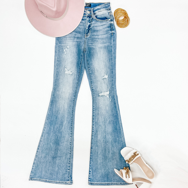 A pair of light wash flare jeans with distressing around the knees. These jeans are pictured on a white background with white heels, a pink hat, and gold jewelry.