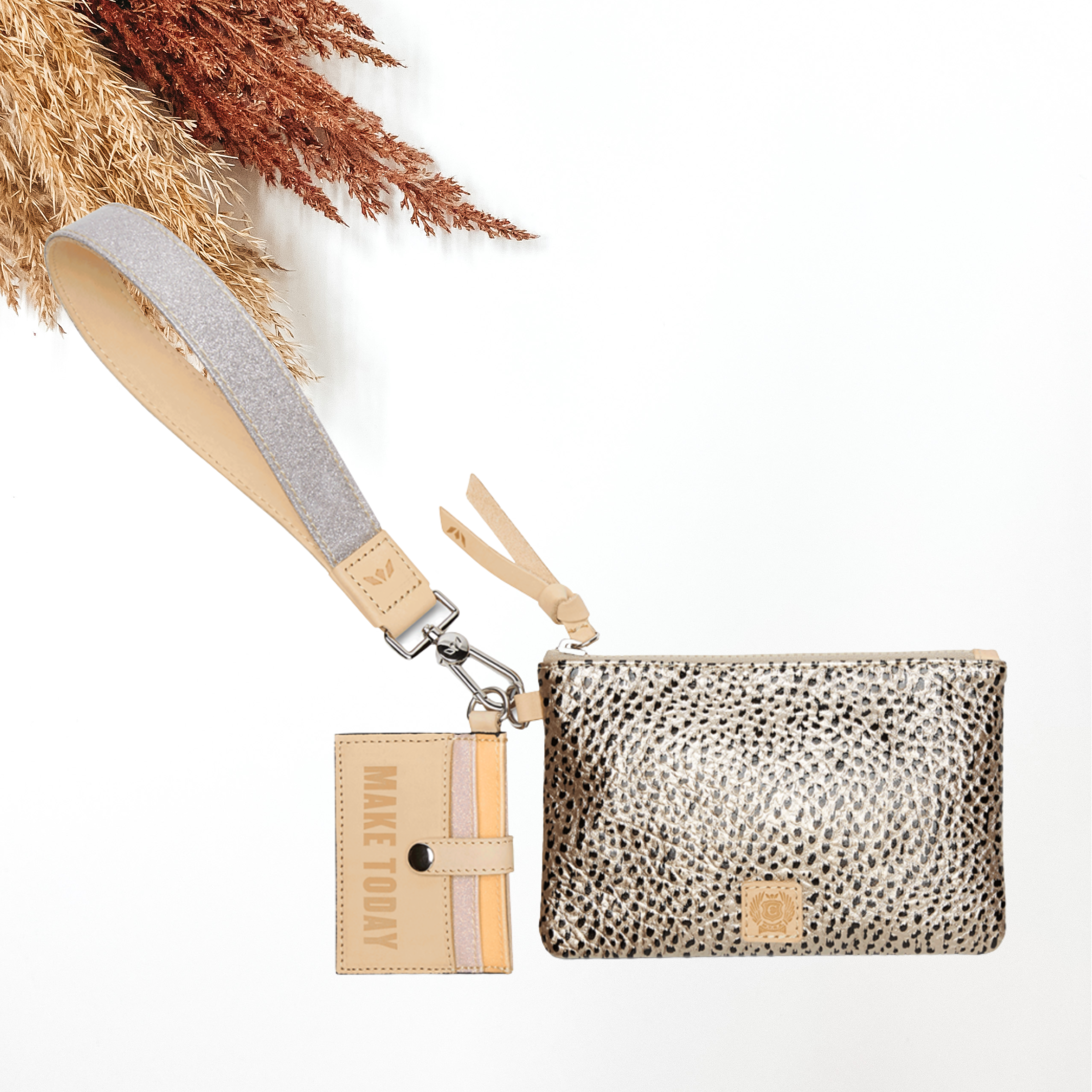 Silver glitter wristlet with a card holder keychain and a tan puch with black dots. This pictured on a white background with tan and brown pompous grass in the top left corner. 