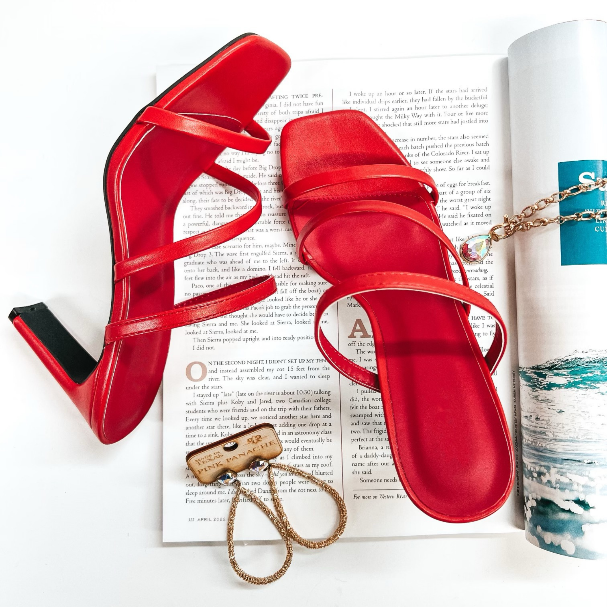 A pair of  red strappy heels with a square toe. These shoes are pictured on a white background with gold jewelry and a magazine.