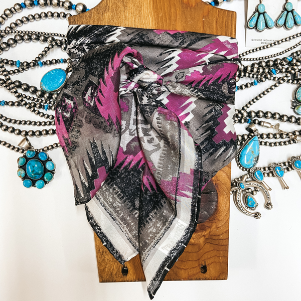 An Aztec print scarf with silver and pink colors. Scarf is tied around a wooden display with turquoise and silver jewelry.
