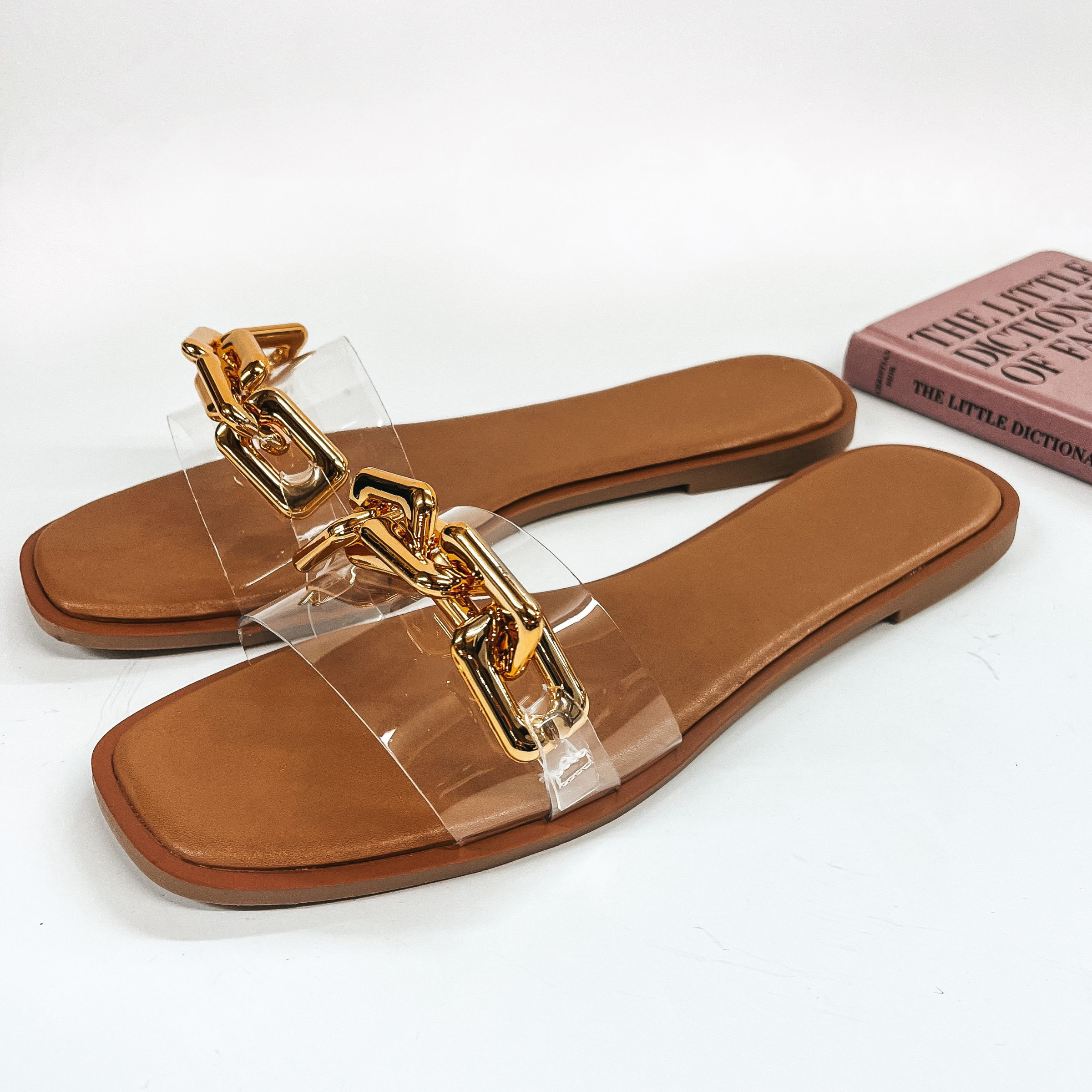 A pair of slide on flat sandals that have a clear strap with a gold chain on top. Pictured on white background with fashion book.