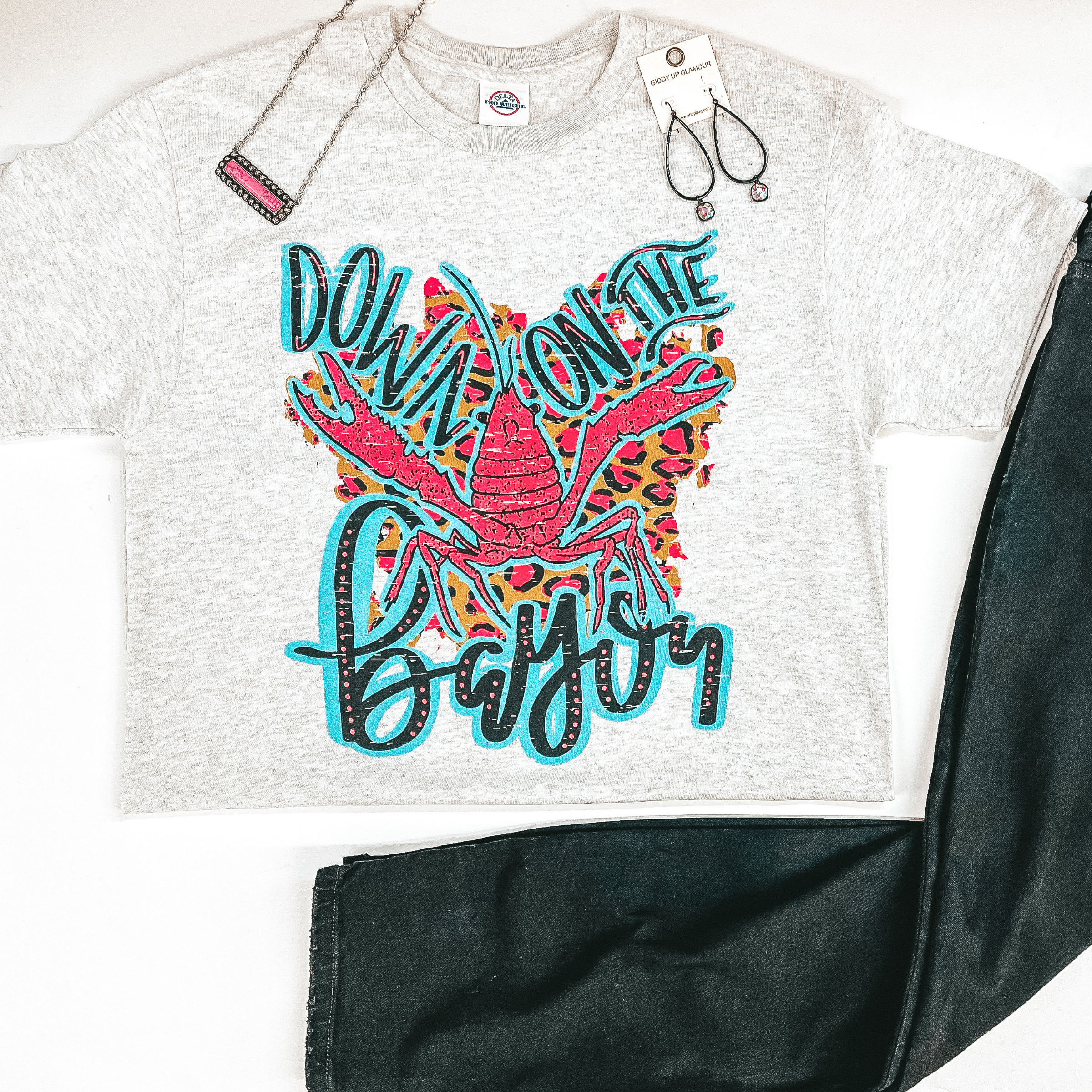 A heather grey tee shirt that is folded in half, exposing the graphic that says "Down On the Bayou" with a hot pink and leopard print crawfish graphic. Pictured with black skinny jeans, hot pink necklace, and crystal earrings.