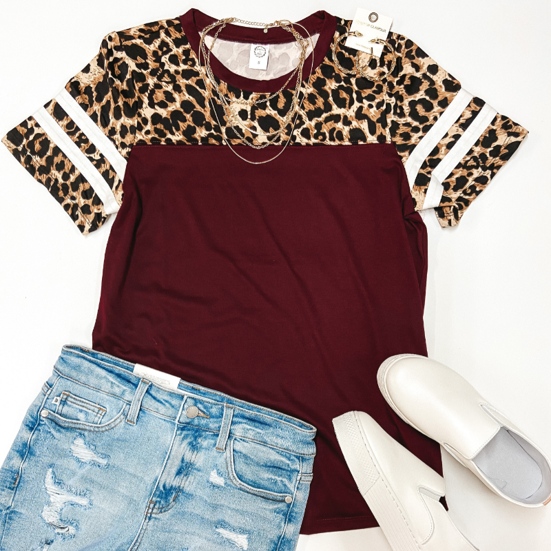 A maroon top with a leopard print short sleeve upper. Pictured with denim shorts, white sneakers, and gold jewelry.