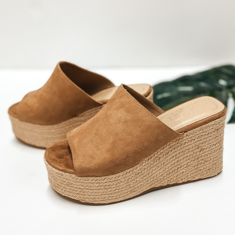 A pair of tan suede slide on wedges that have a rope sole. Pictured on white background with a palm leaf.