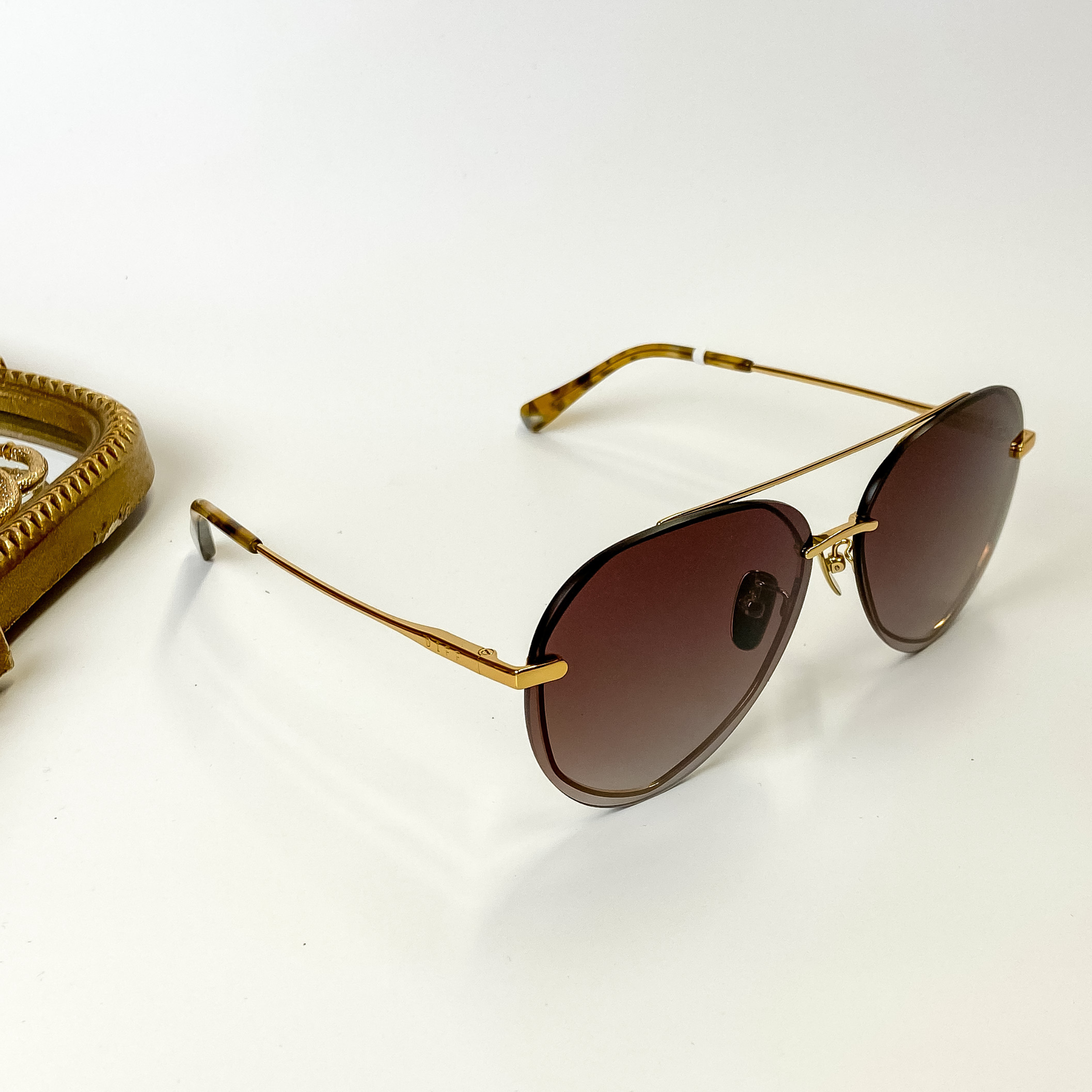 A pair of gold-tone aviator style glasses with brown lenses. These sunglasses are pictured on a white background with gold jewelry.