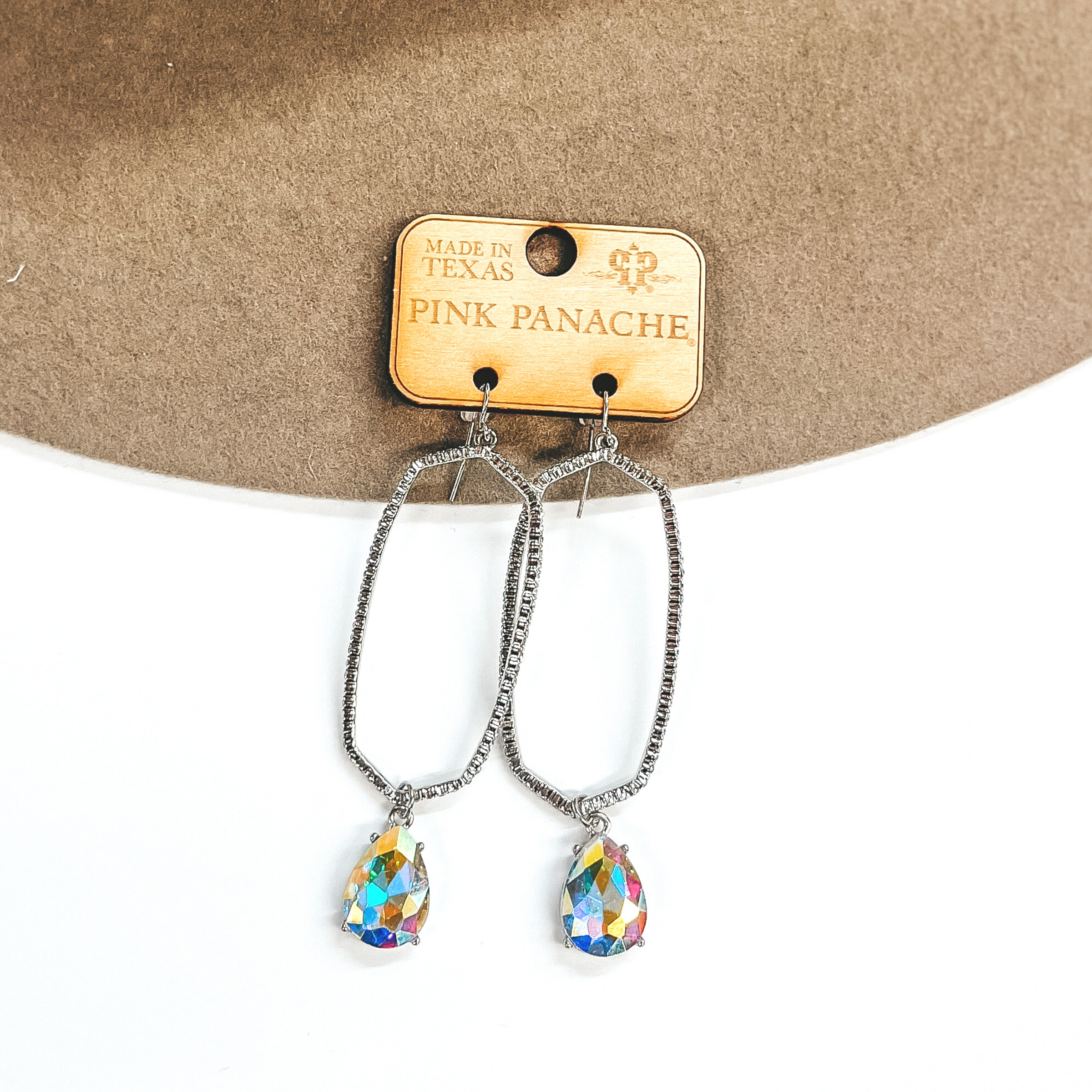 A pair of oval outline earrings that have crystals all the way around. These earrings have an AB crystal teardrop pendant at the bottom of each earring. Pictured on white background with a beige hat. 