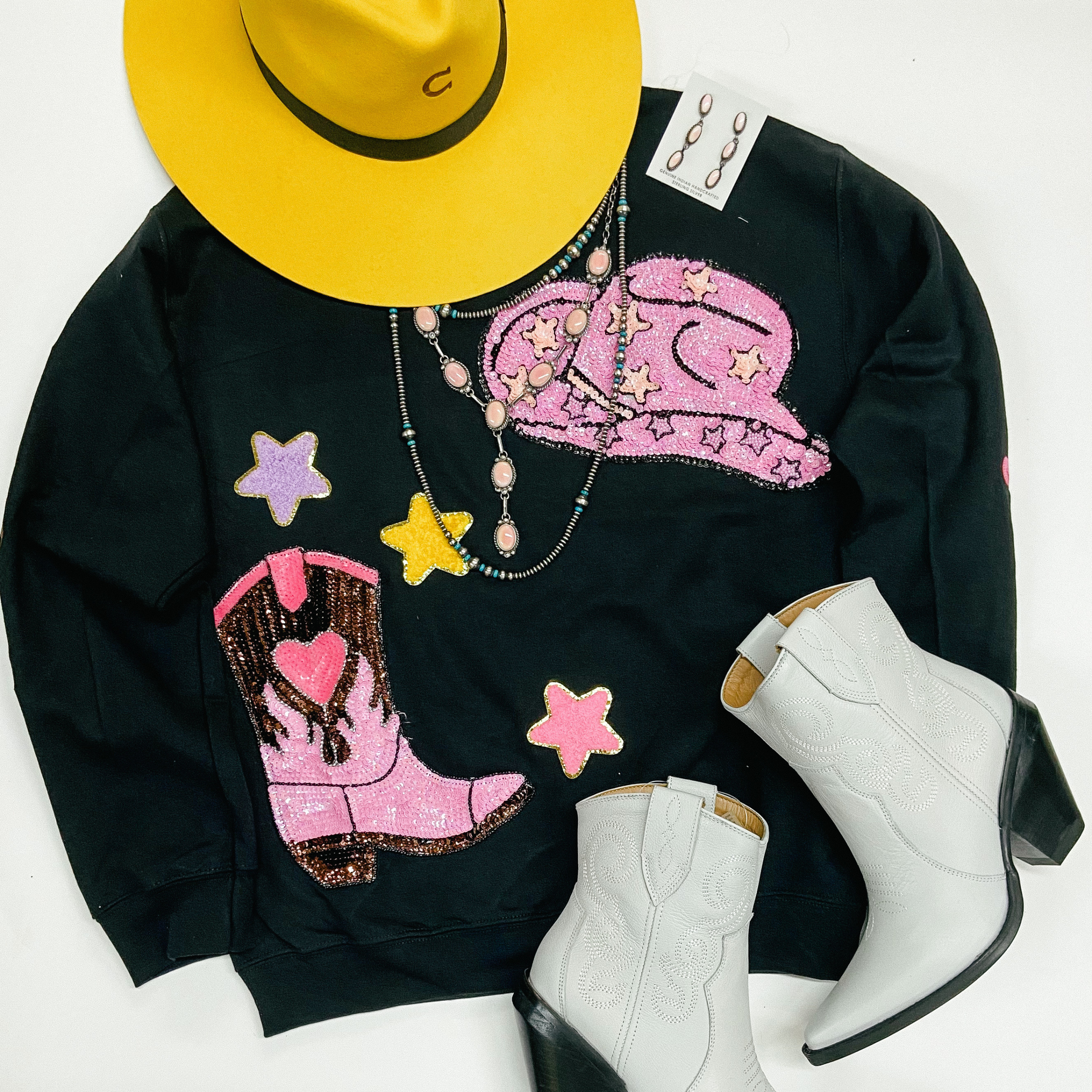 A black sweatshirt with a pink sequin cowboy boot patch, pink cowboy hat patch, and chenille star patches. This sweatshirt is pictured on a white background with a yellow cowboy hat, white booties, and genuine Navajo handmade jewelry.