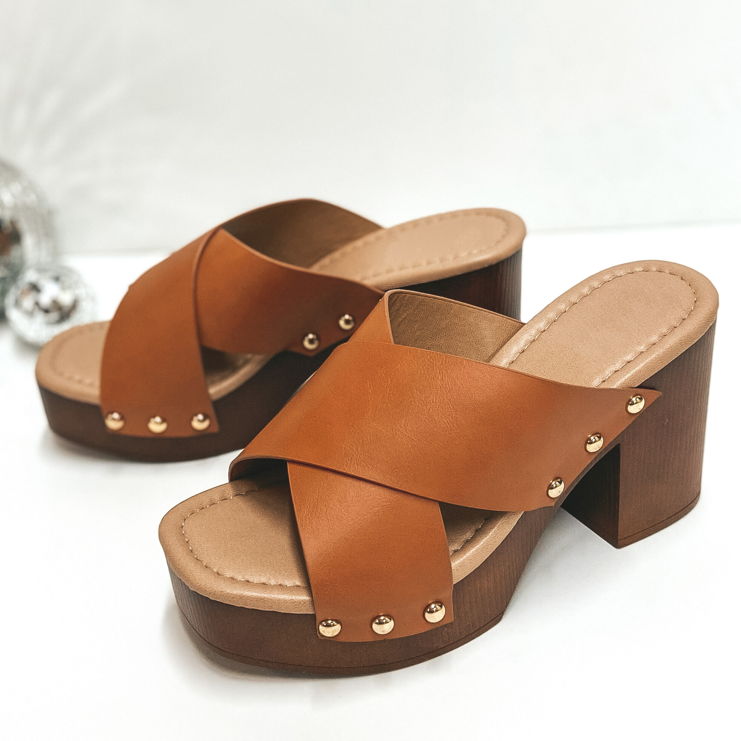 A pair of clog style heels with a cognac brown criss cross band. Pictured on white background with disco balls.
