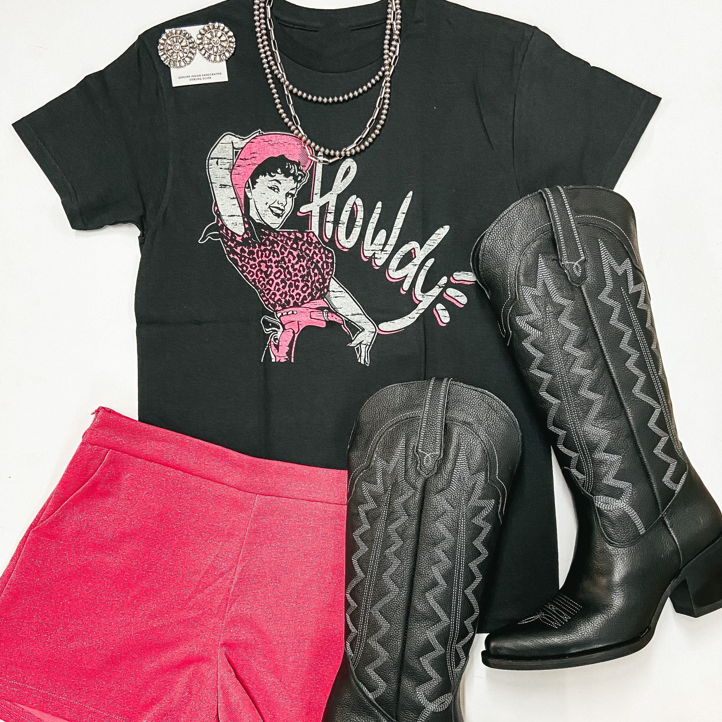 The graphic tee is black and says "Howdy" with a cowgirl wearing pink. The tee shirt is pictured with a black hat, pink sequin shorts, black cowgirl boots, and sterling silver Navajo Pearls.