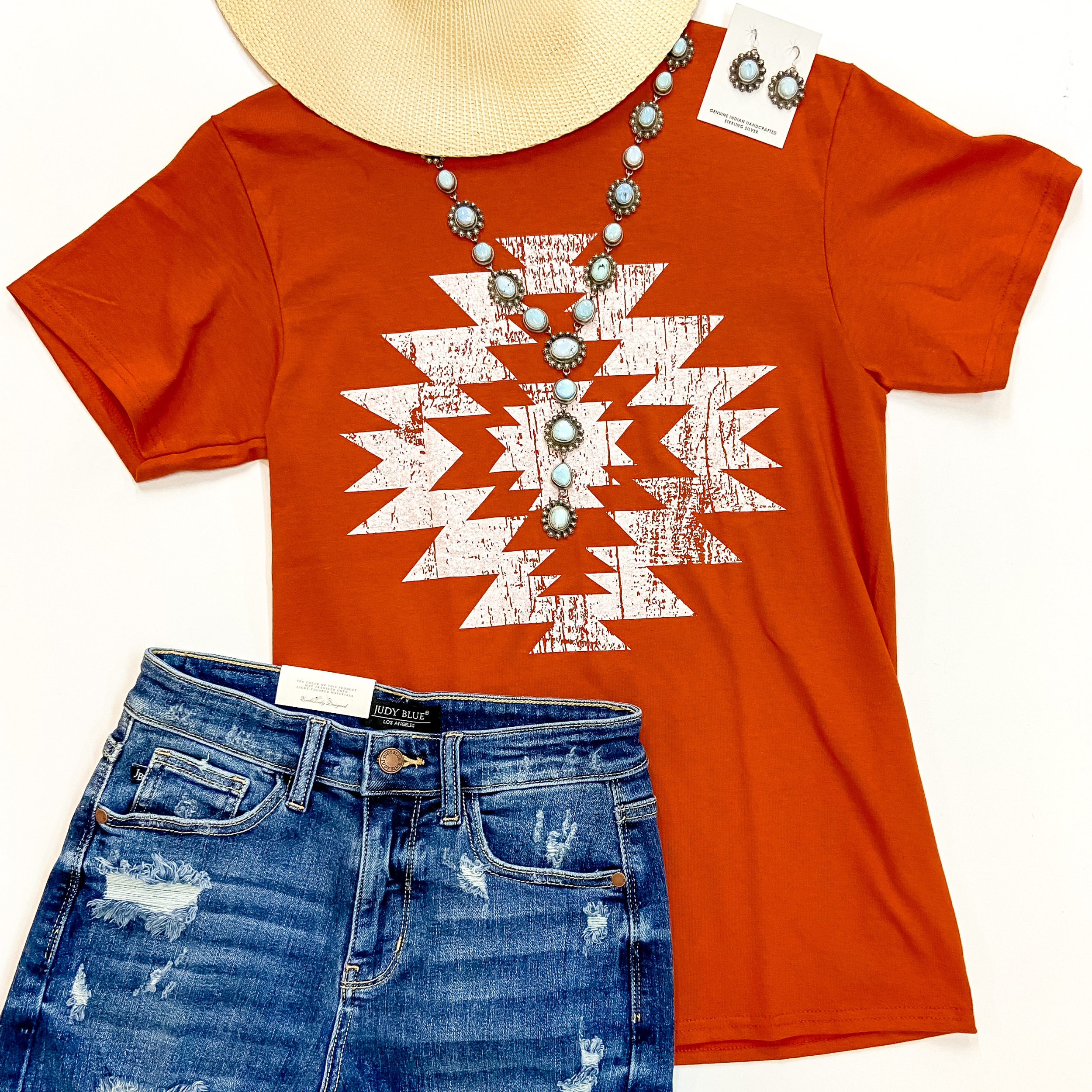 A burnt orange short sleeve tee shirt with a crew neckline and a graphic of a tribal symbol. This tee shirt is pictured on a white background with denim shorts, genuine sterling silver and turquoise jewelry, and a straw hat.
