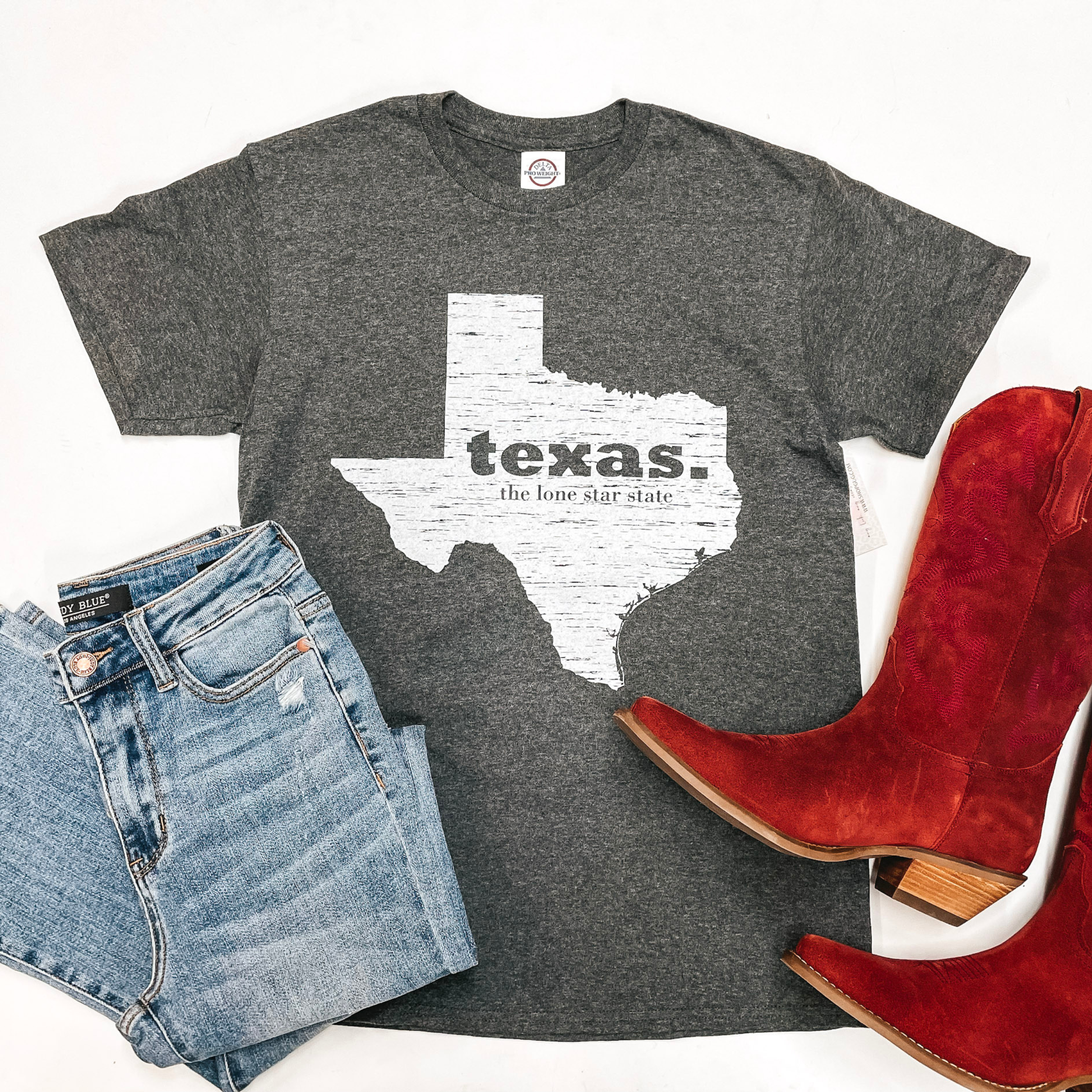 A gray tee is in the middle of the picture on a white background. In the center of the tee is a Texas with block letters saying "texas. the lone star state'" On the bottom left of the picture is light-wash jeans and on tthe bottom right of the picture is red boots. 