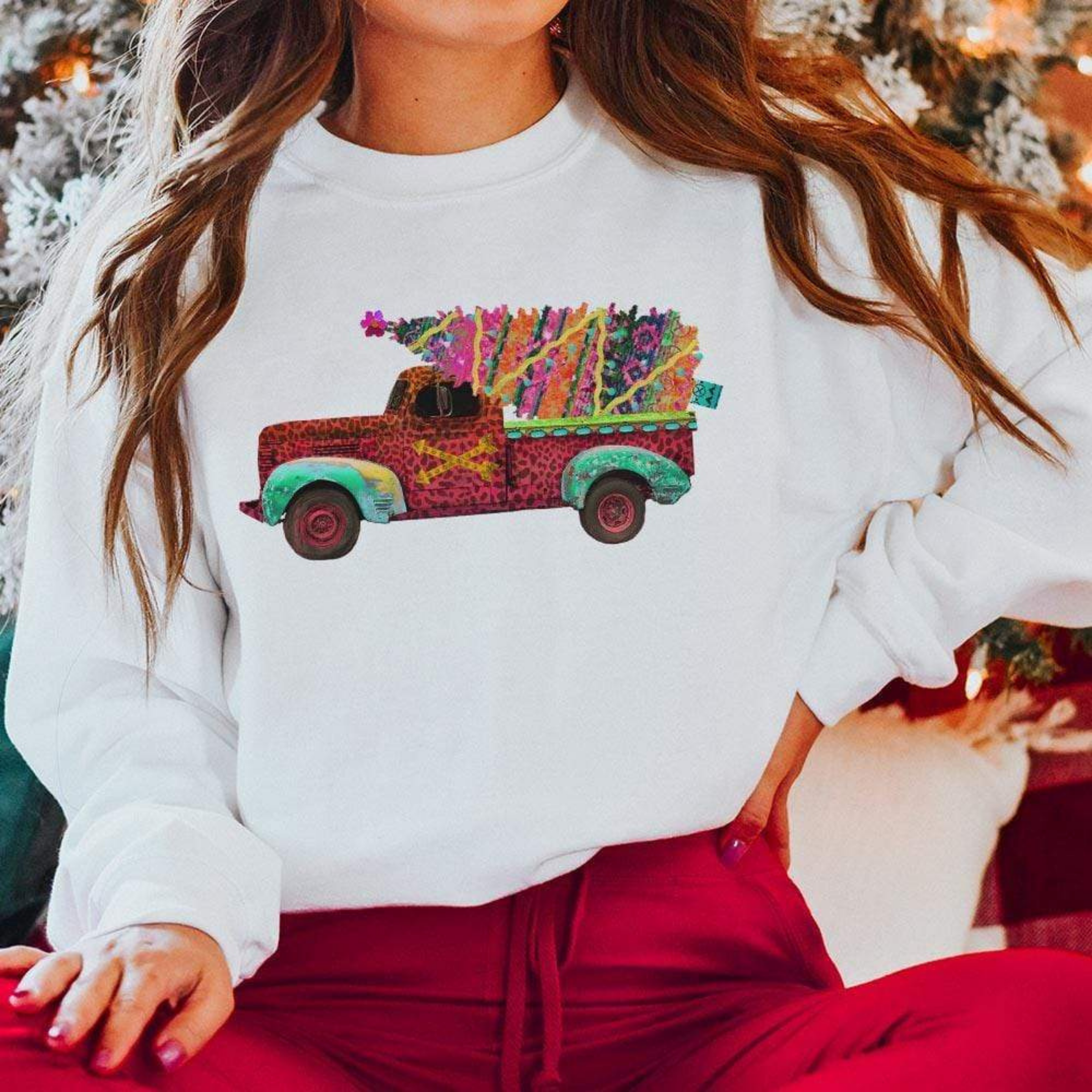Model is wearing a white sweatshirt with long sleeves and a graphic of a leopard pick up truck, hauling a serape christmas tree. Model has it paired with red pants and is sitting infront of a christmas tree.