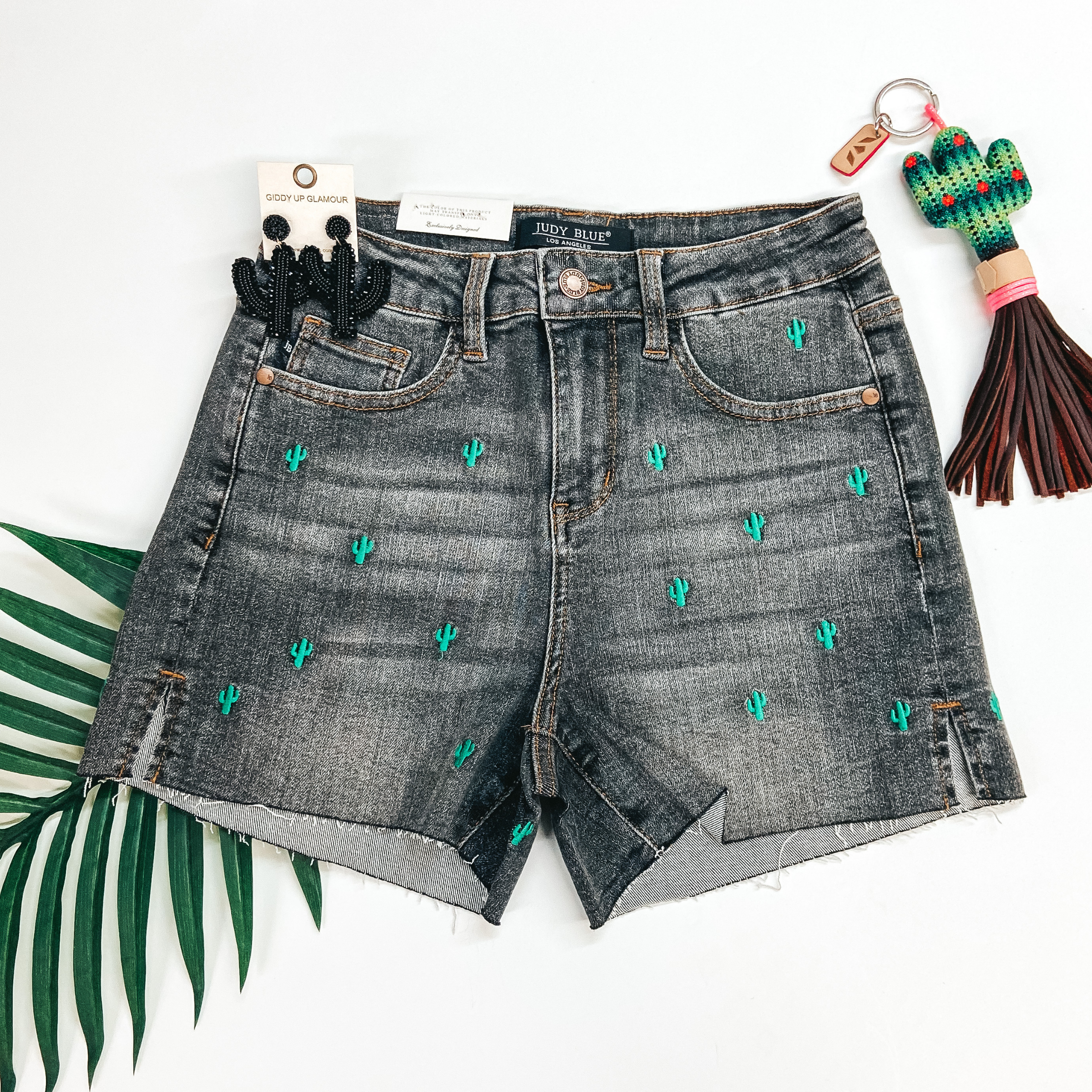 A pair of black washed shorts that are cut off. These shorts have tiny saguaro cacti embroidered all over. Shorts are pictured on a white background with cactus earrings, a cactus keychain, and a palm leaf.