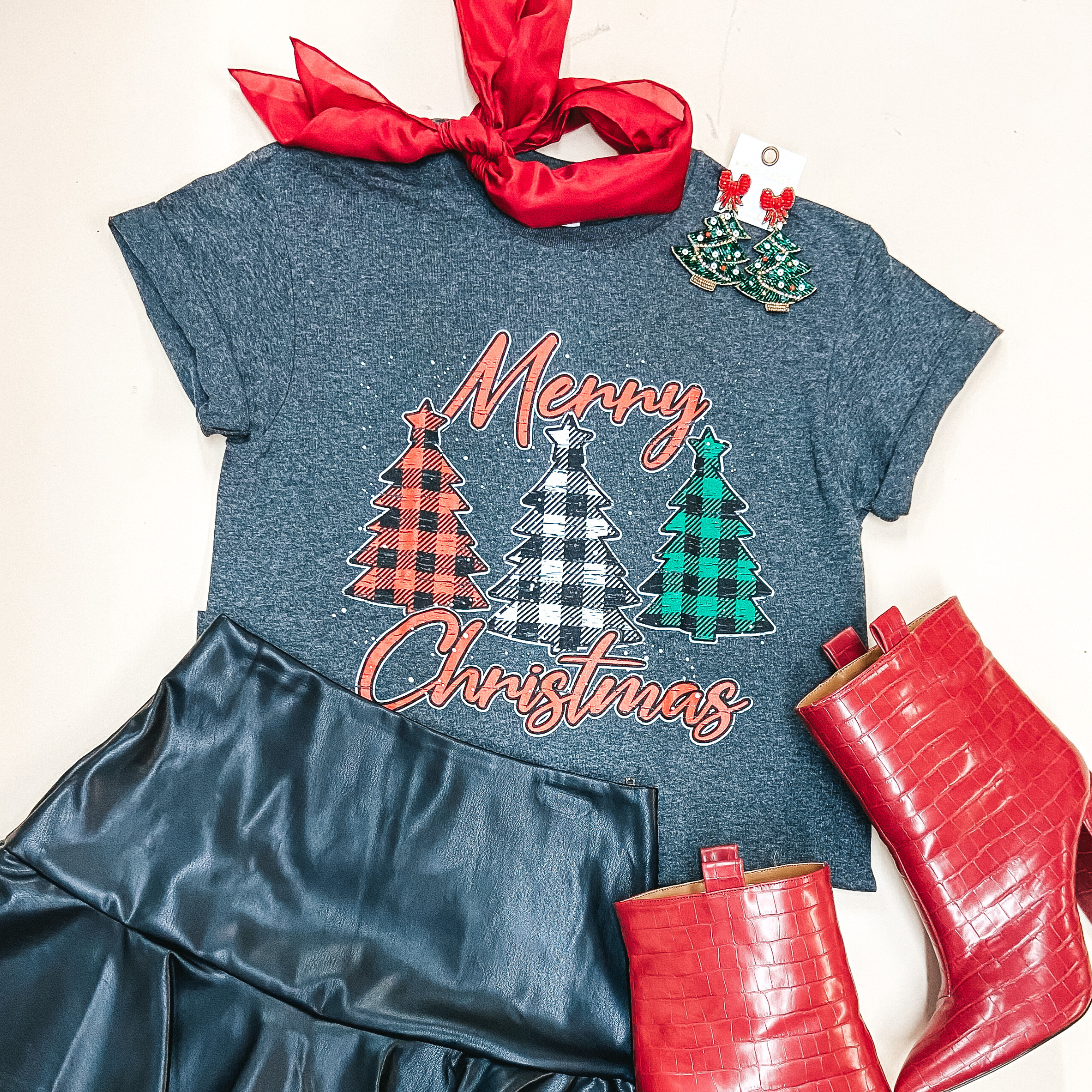 A heather grey short sleeve tee shirt with a plaid Christmas tree graphic that says "Merry Christmas" in red letters. Pictured with red booties, a red wild rag, and a black faux leather skirt.