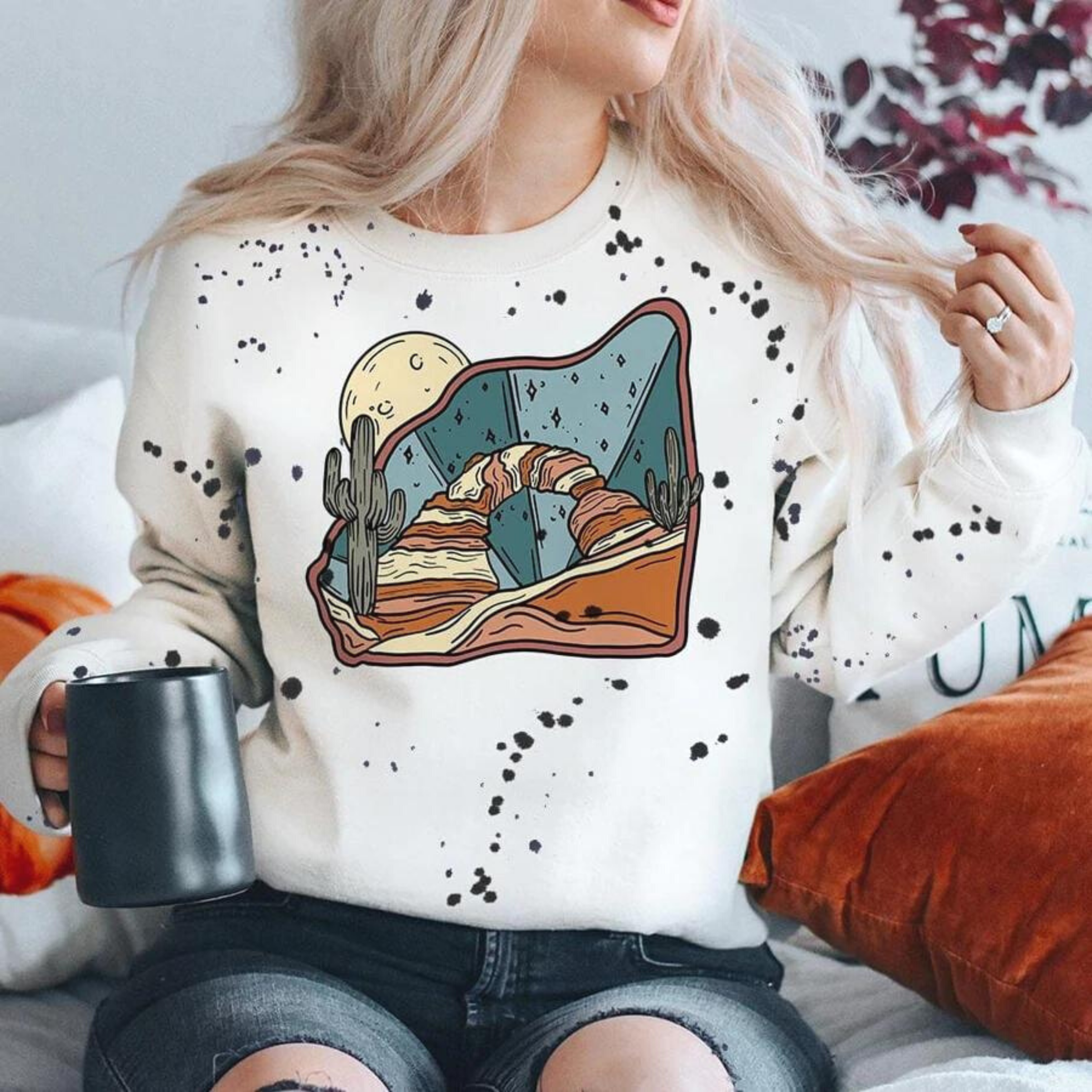 A long sleeve graphic sweatshirt that has an image of a rock arch with saguaro cactus and a moon on a splatter paint background.