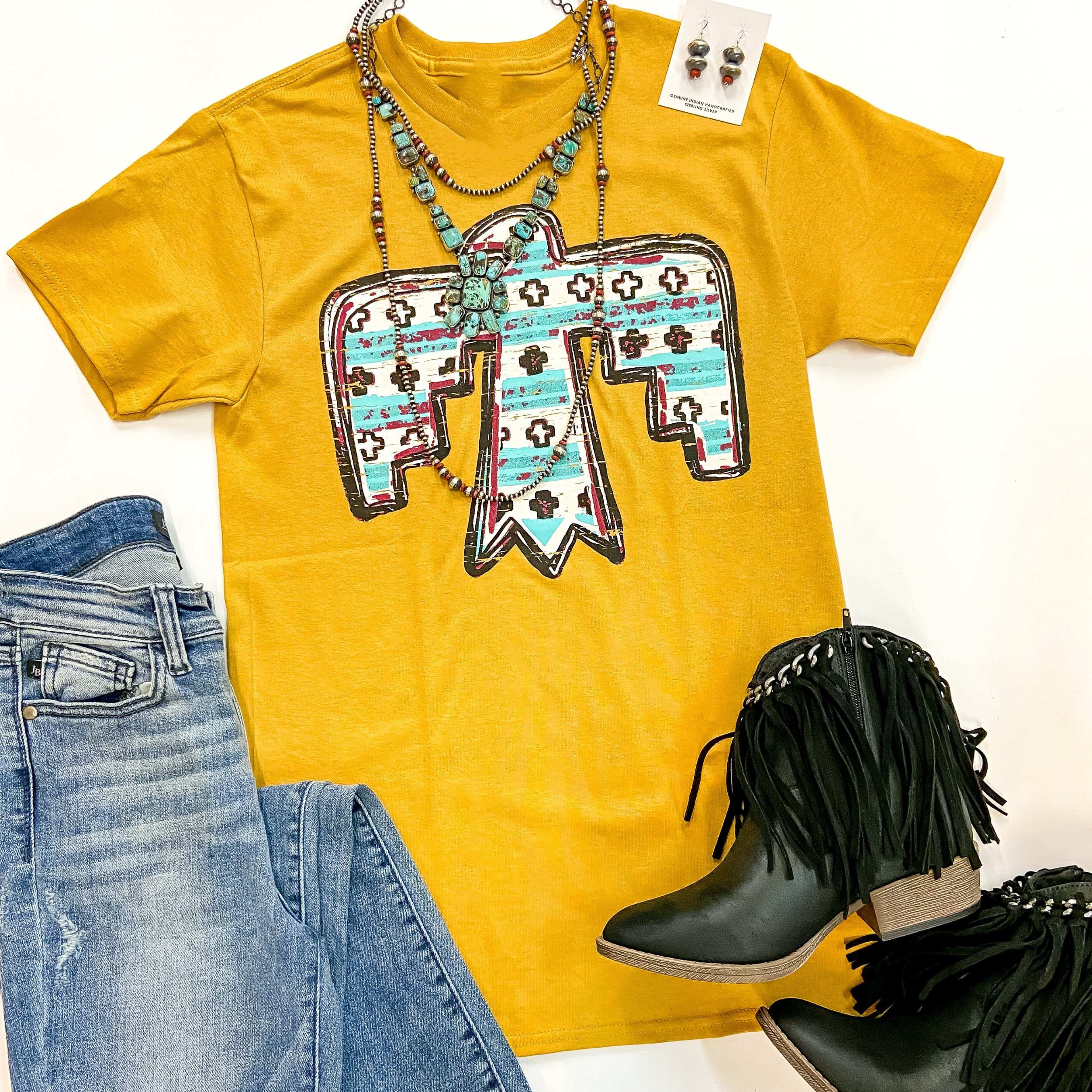 A mustard yellow short sleeve tee shirt with a crew neckline and a graphic of a thunderbird with ivory, turquoise, and red tribal print on the thunderbird. This tee is pictured with denim jeans, black booties, and genuine turquoise and sterling silver jewelry.