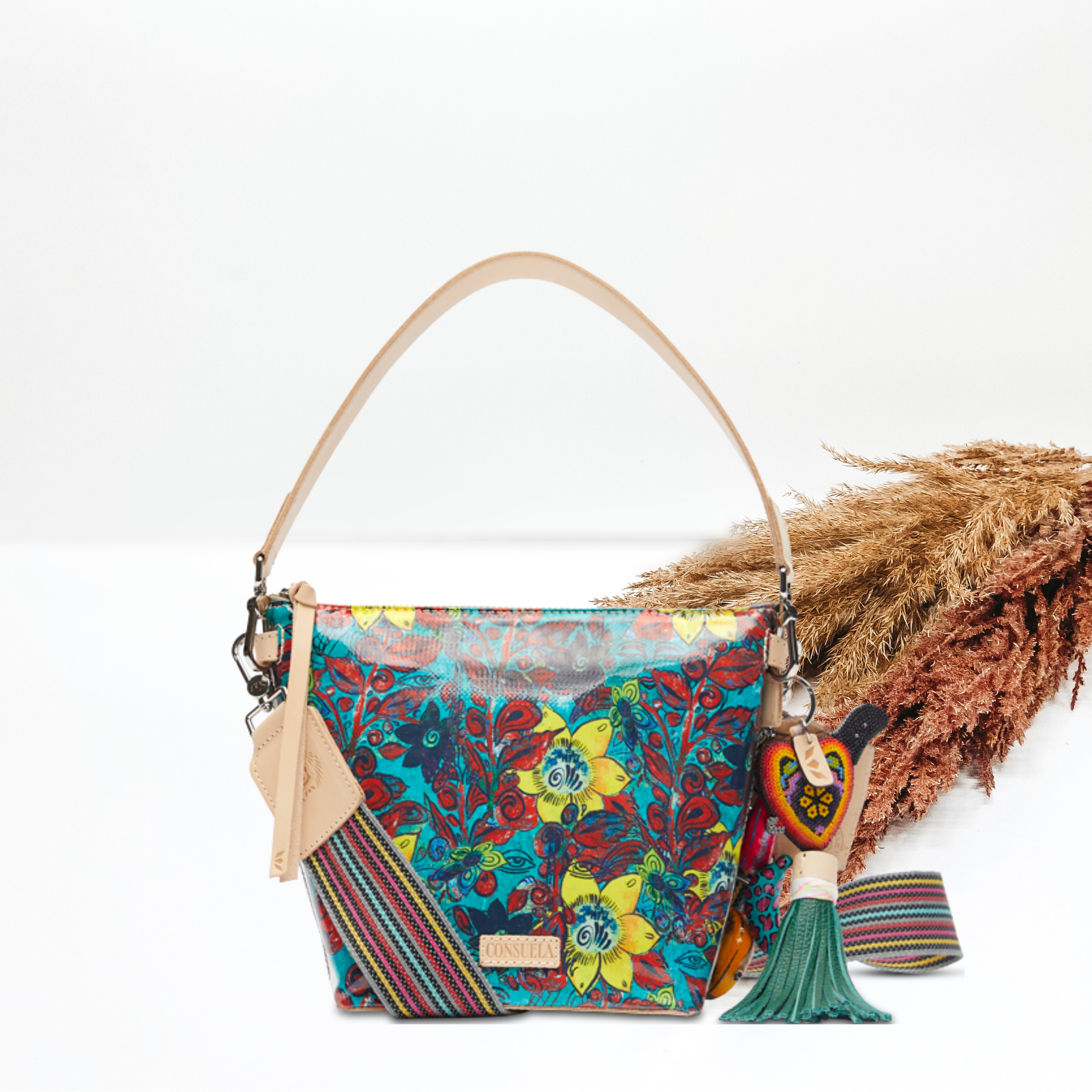 In the middle of the picture is a wedge bag in a blue, yellow, and red floral print. The background is solid white with pompas grass to the right of the bag.