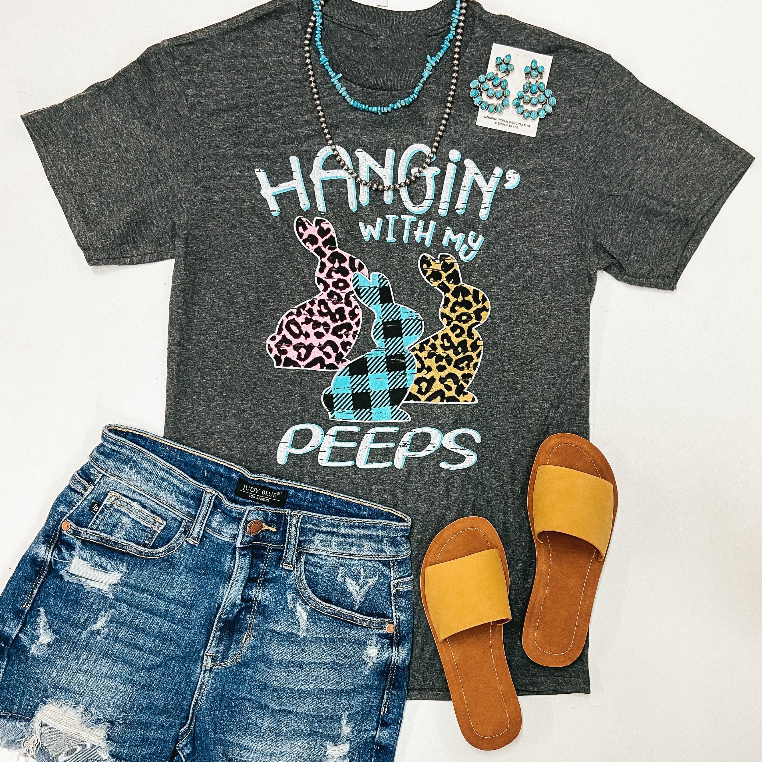 A charcoal grey tee shirt with cuffed sleeves and knotted hemline. The tee shirt has a graphic that says "Hangin' with my peeps." and three bunnies that are pink leopard print, blue buffalo plaid print, and yellow leopard print. Tee shirt is pictured on white background with denim shorts, a mustard yellow hat and sandals, and silver jewelry.