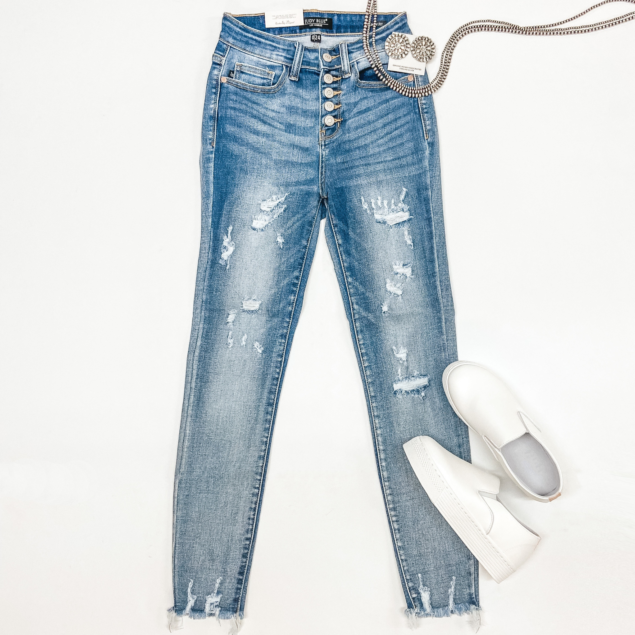 A pair of button fly distressed jeans with a raw hem. These jeans are pictured on a white background with sterling silver jewelry and white sneakers.