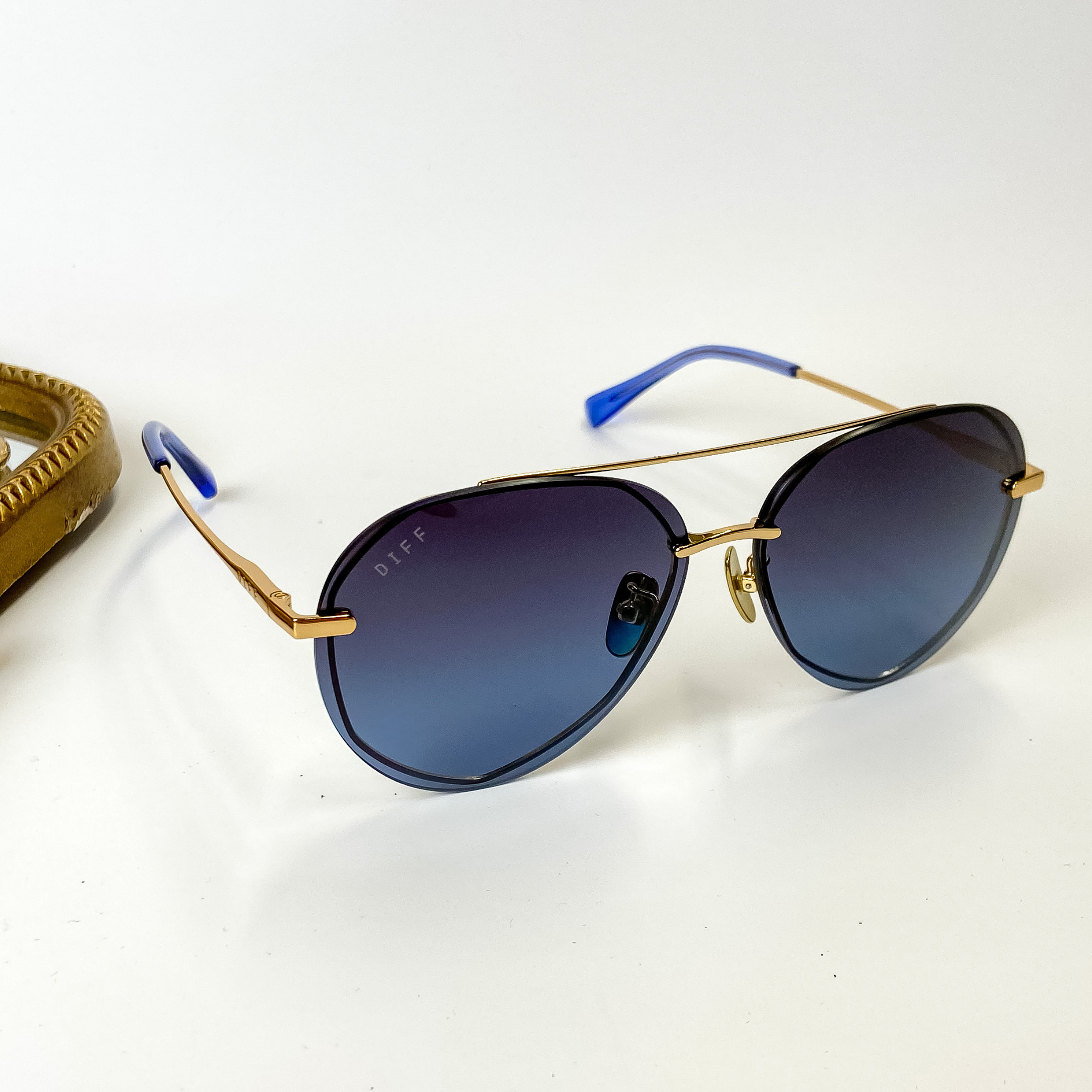 A pair of gold tone aviator style sunglasses with blue lenses. Pictured on a white background with gold jewelry.