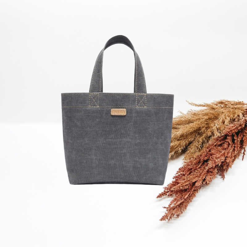 Centered in the picture is a small tote bag in a soft blue material. Background is solid white with pampas grass laid to the right of the bag. 