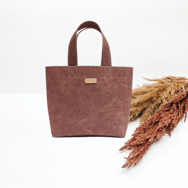 Pictured is a small tote bag in a brown-mauve color. Background is solid white with pompas grass laying to the right of the bag.  