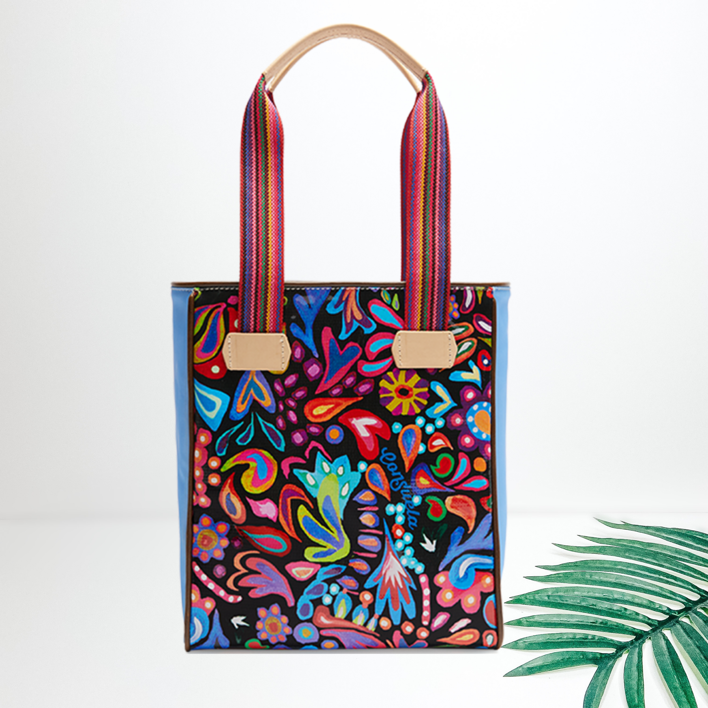 A black shoulder bag with a multi color pattern. Pictured on a white background with a palm leaf.