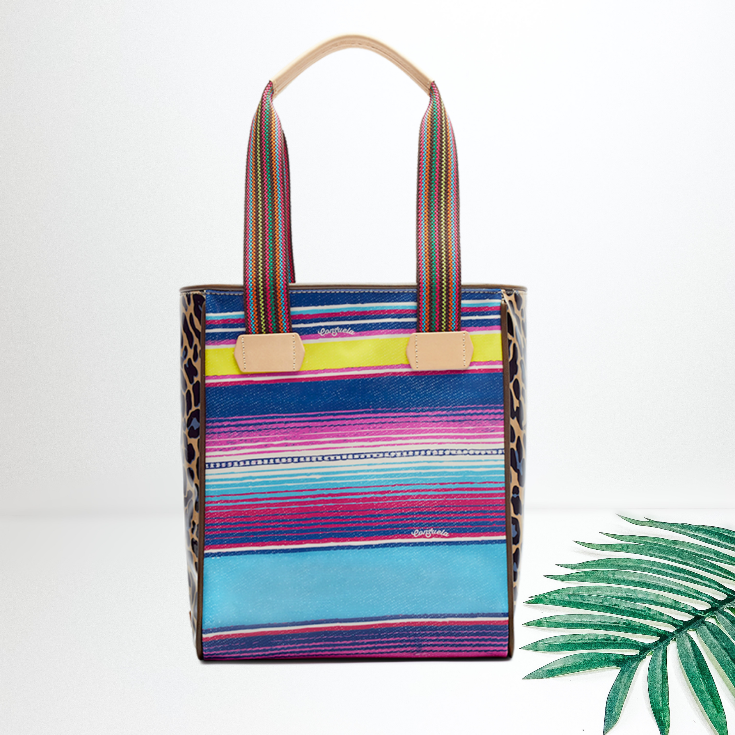 Centered in the middle of the picture is a tote bag in a multi-colored serape print. To the right of the bag is a palm leaf, all on a white background. 