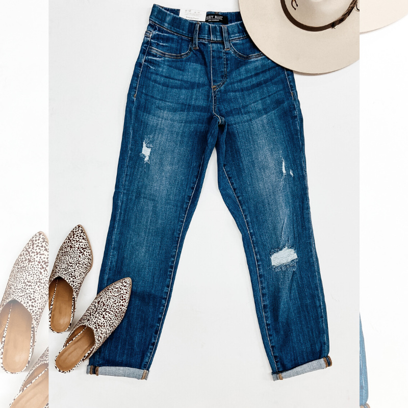 A pair of elastic waist skinny jeans pictured on a white background with a beige hat and dotted mules.