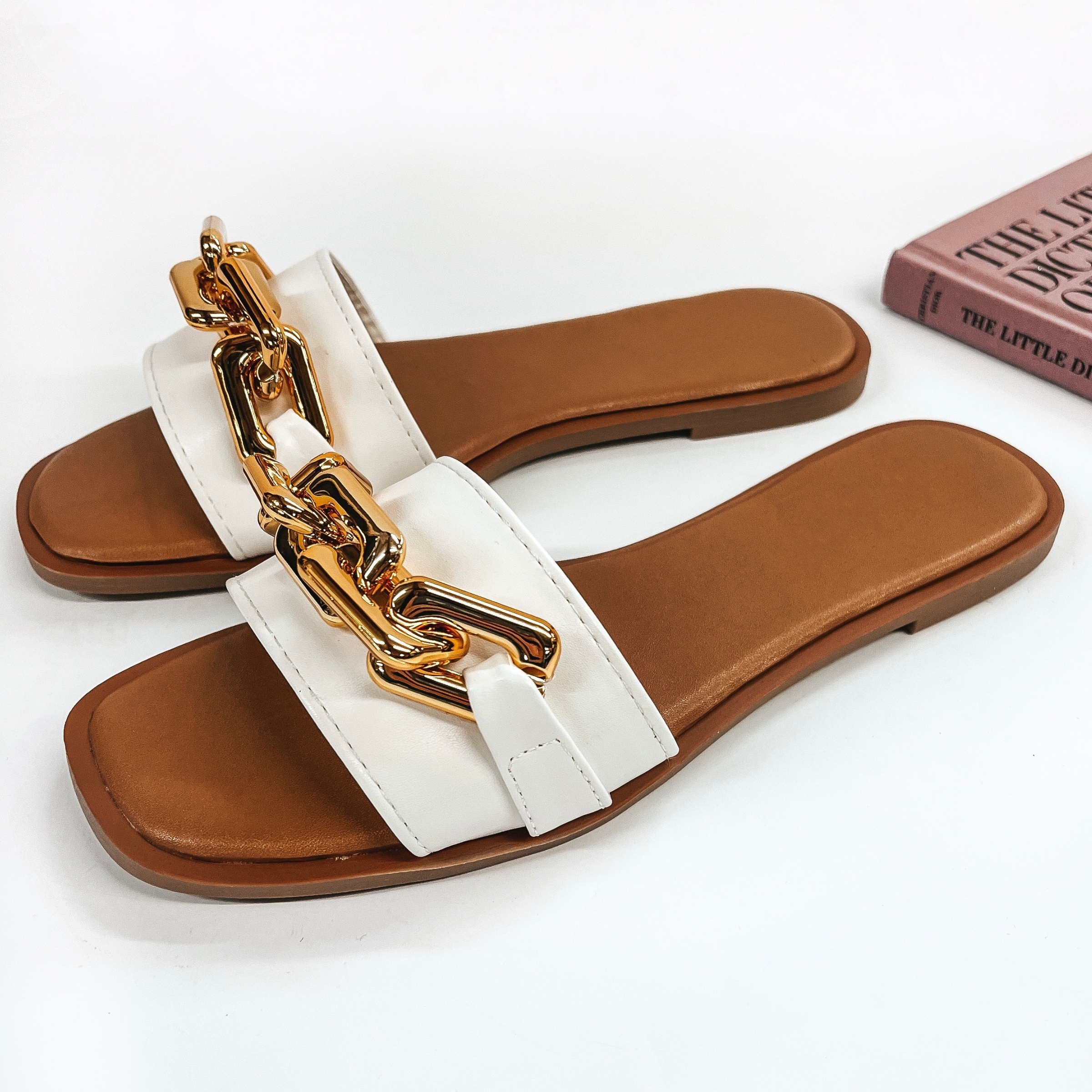 A pair of slide on flat sandals that have a white strap with a gold chain on top. Pictured on white background with fashion book.