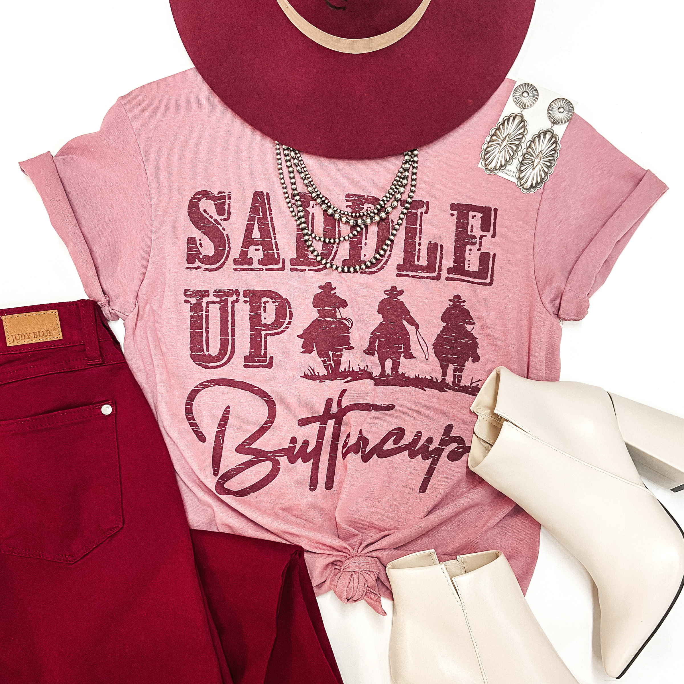 A mauve pink tee shirt with cuffed sleeves and a knotted front. The tee shirt has a maroon graphic of cowboys on horses that says "Saddle Up Buttercup." Pictured on white background with a maroon hat, maroon jeans, and sterling silver jewelry.
