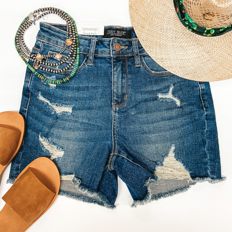 A pair of distressed dark wash shorts. Pictured on white background with tan sandals, a straw hat, and Navajo pearls.