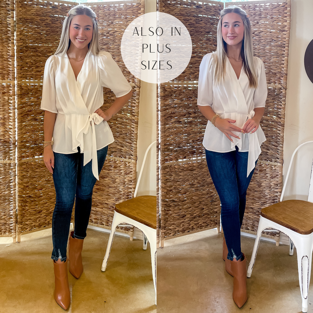 Model is wearing a white wrap top with a tie. Model has this top paired with dark wash skinny jeans, tan booties, and gold jewelry.