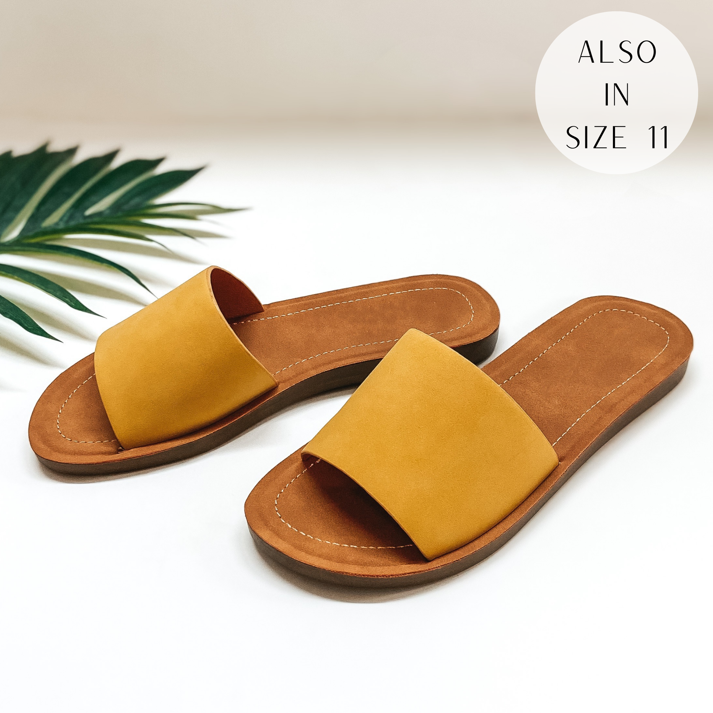 A pair of yellow slide on sandals pictured on a white background with a palm leaf.