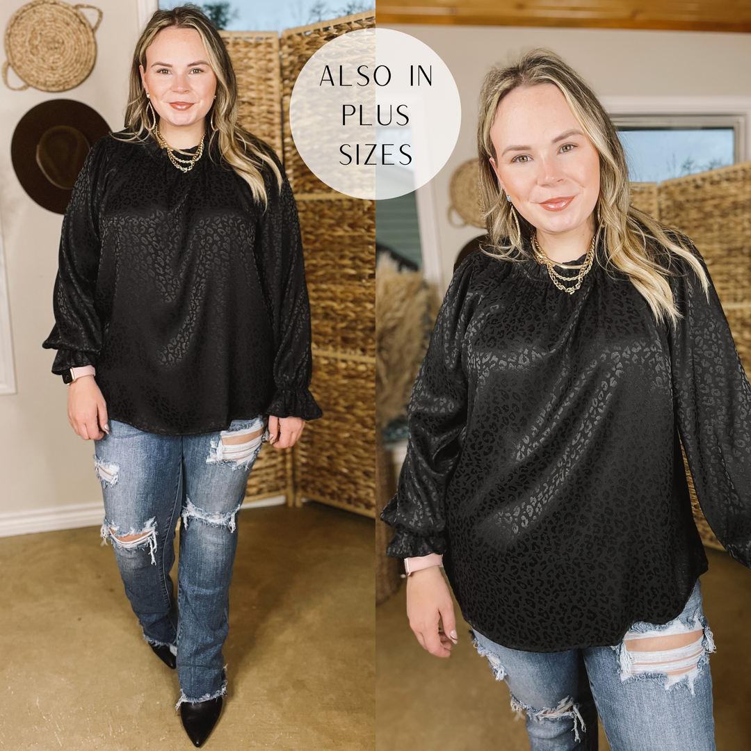 Model is wearing a black satin long sleeve shirt in a leopard print with a ruffled detail around the wrist. Model has this top paired with distressed jeans and black booties with a gold necklace and earrings.  Background is brown. 